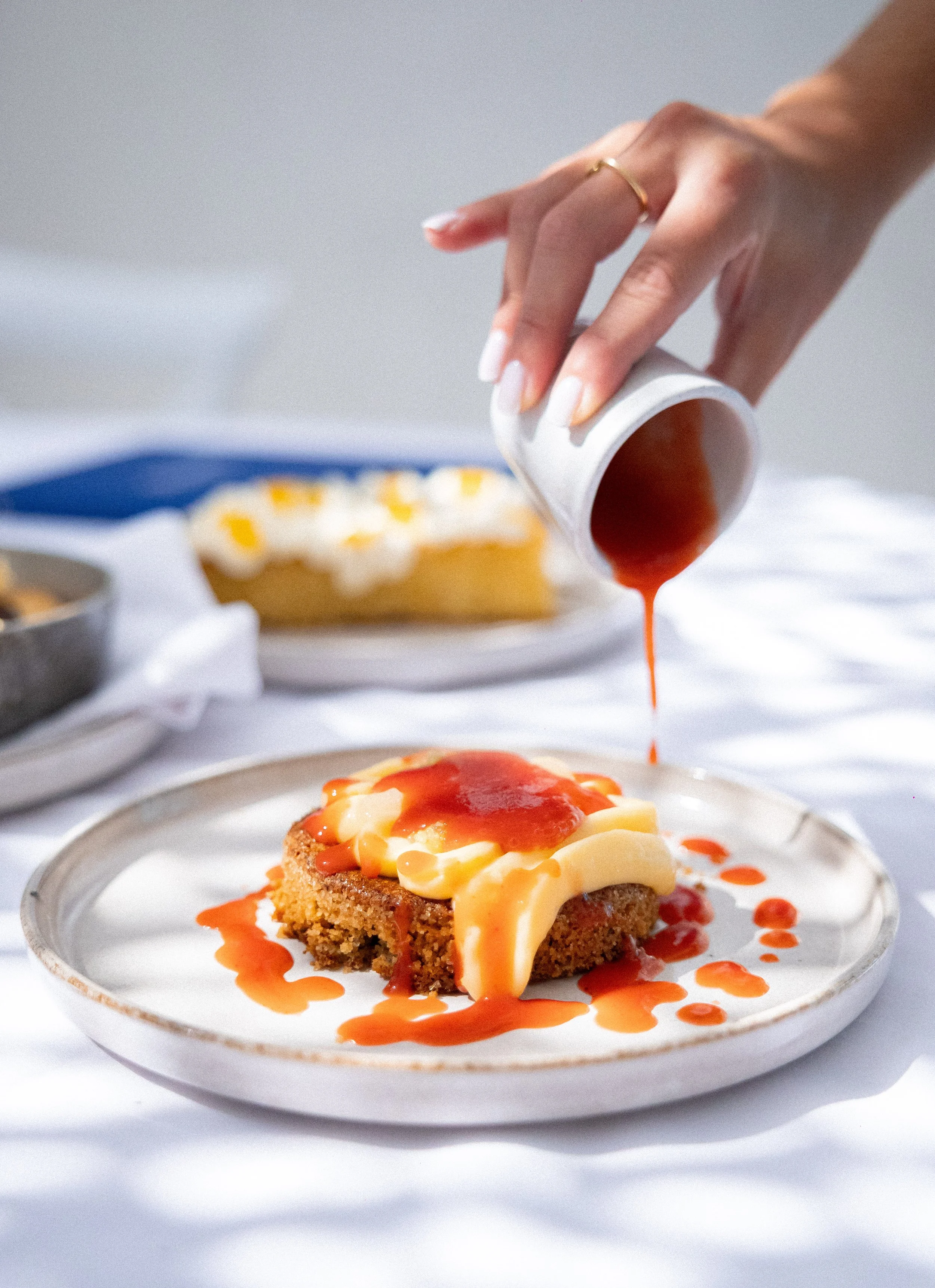 A dessert on a white plate with a piece of cake topped with cream, orange sauce, and yellow-colored garnish. A hand is pouring red sauce over the dessert from a small white cup.