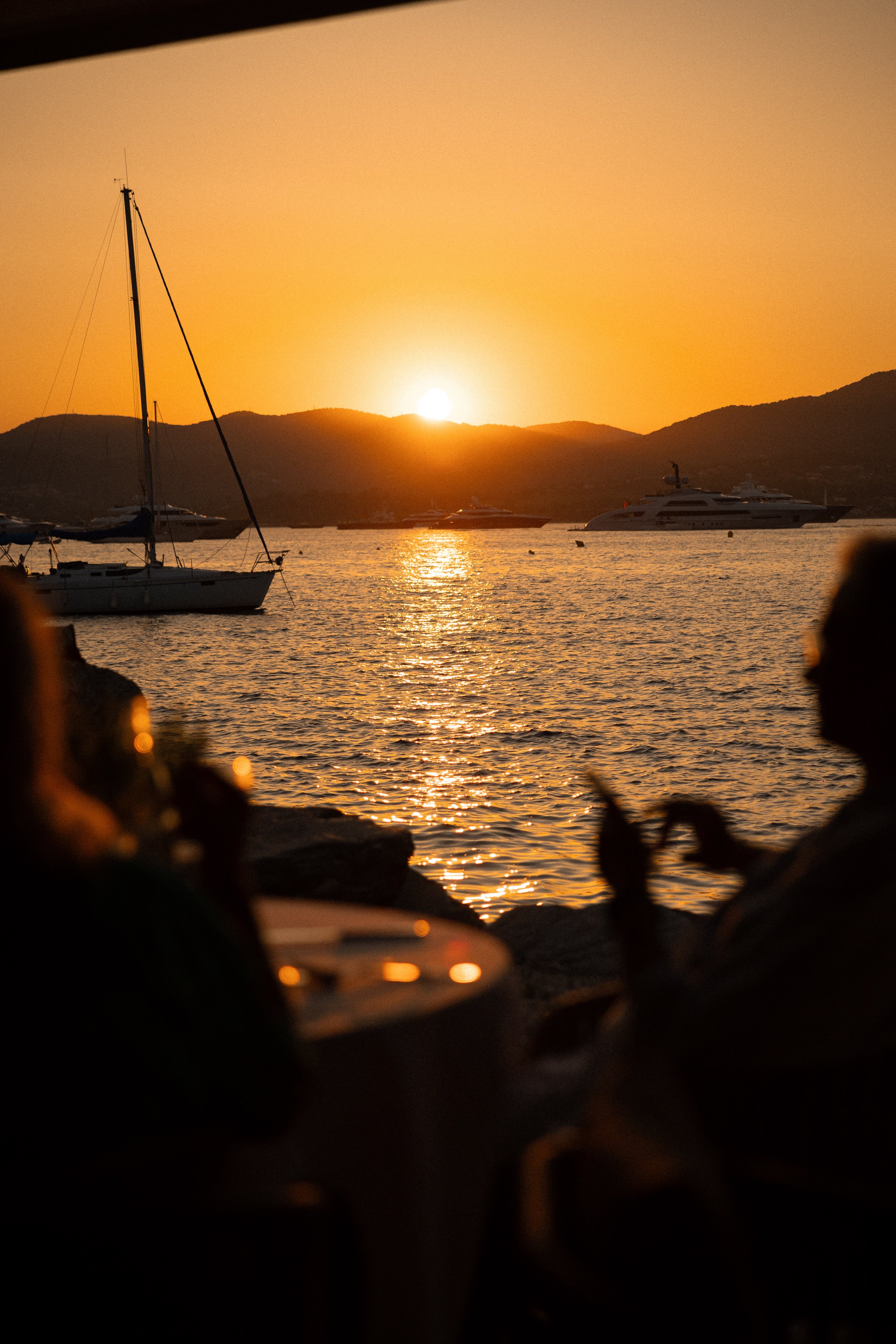 Sunset over the water with sailboats and a yacht, mountains in the background, viewed from a rocky foreground.