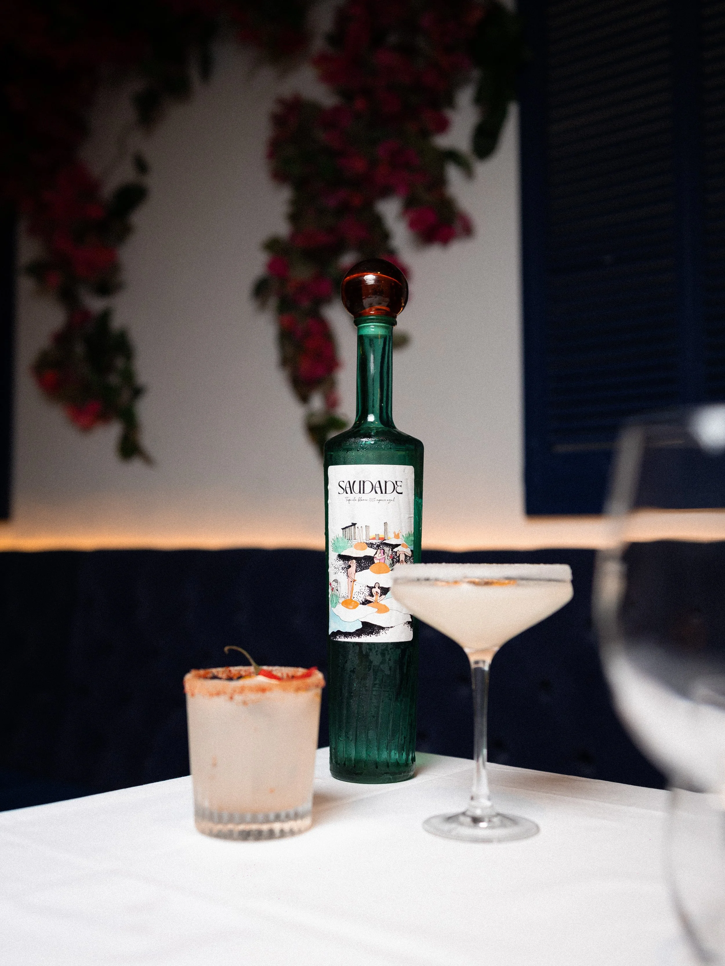 A small green glass bottle with a colorful label featuring illustrations of people on a white surface, with a white dessert in a glass cup in the foreground, and blurred flowers in the background.