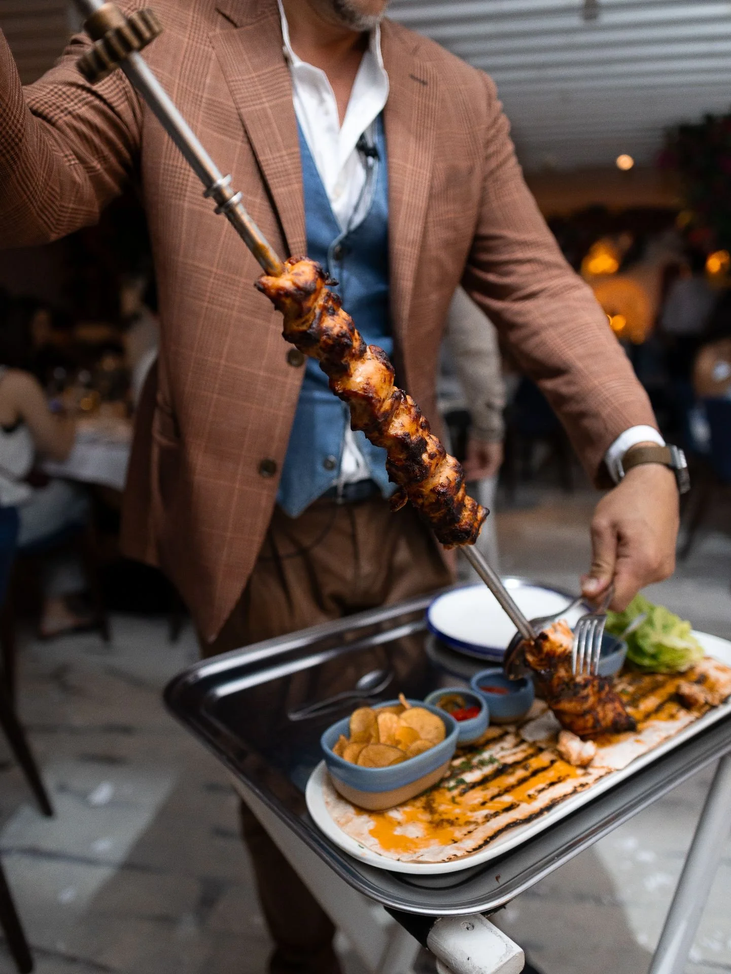 Man in brown checkered suit serving grilled meat on a skewer at a party or gathering with food on a tray.