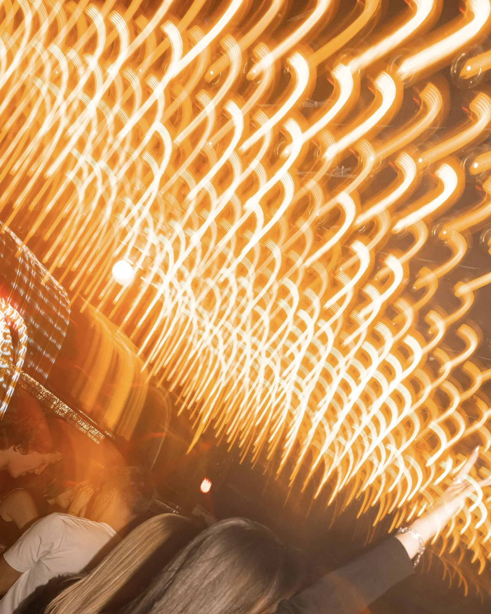 People sitting in a dimly lit area with bright, colored, diagonal, glowing lights overhead, creating a lively and energetic atmosphere.