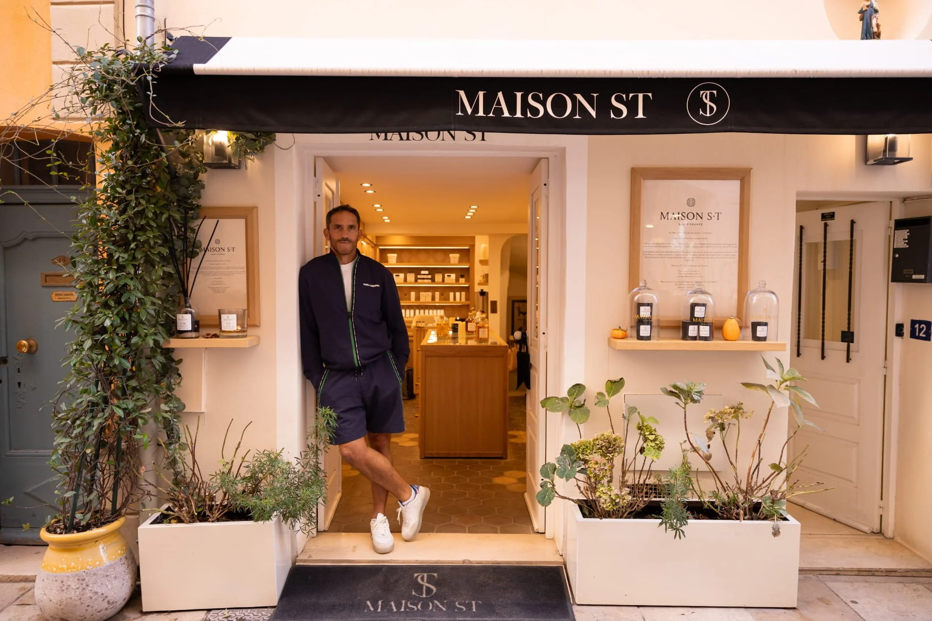 A man standing at the entrance of a shop called MAISON ST, with a black awning displaying the shop's name, surrounded by potted plants and framed posters on the wall inside.
