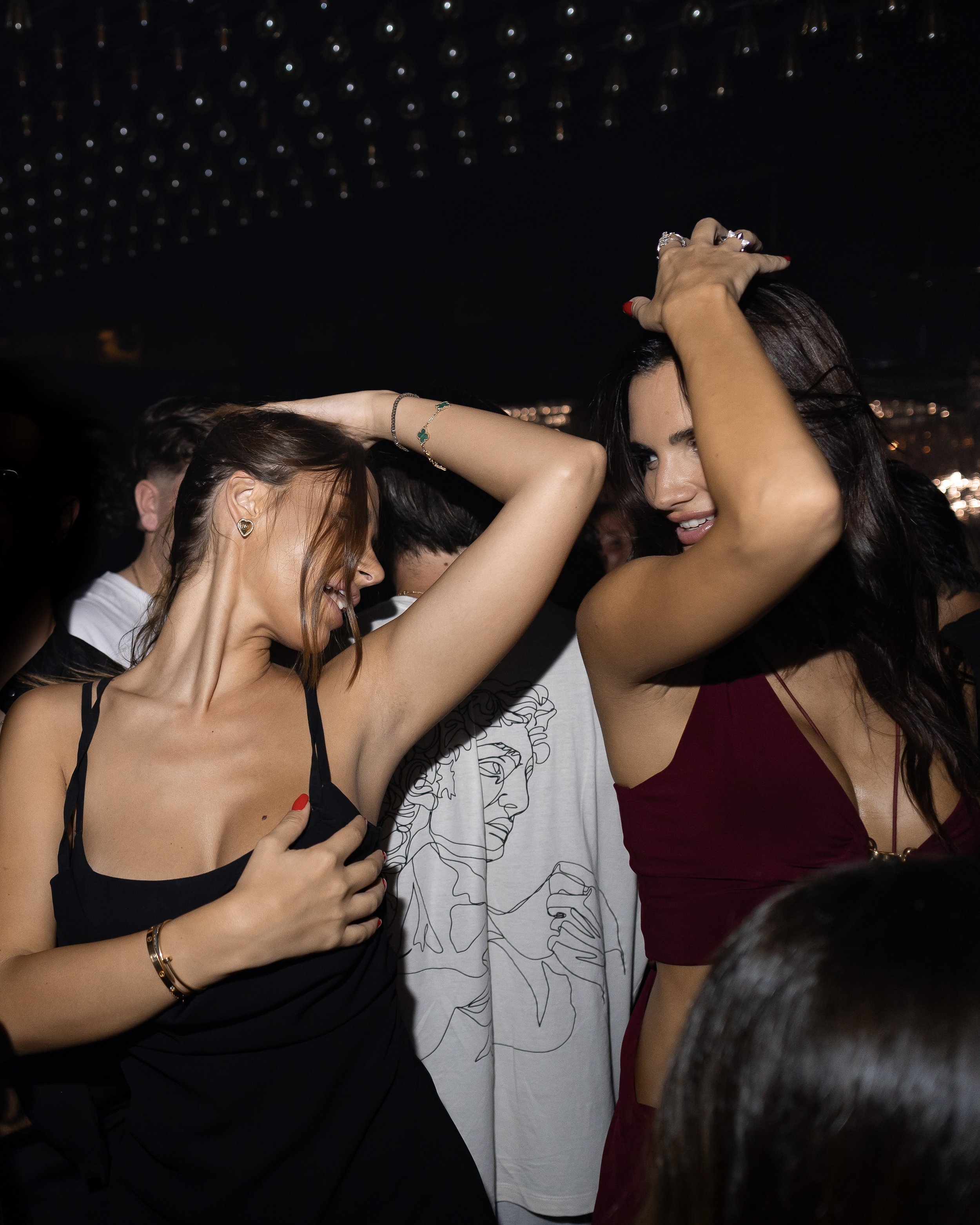 A group of women dancing closely at a party or club, with dark background and dim lighting.