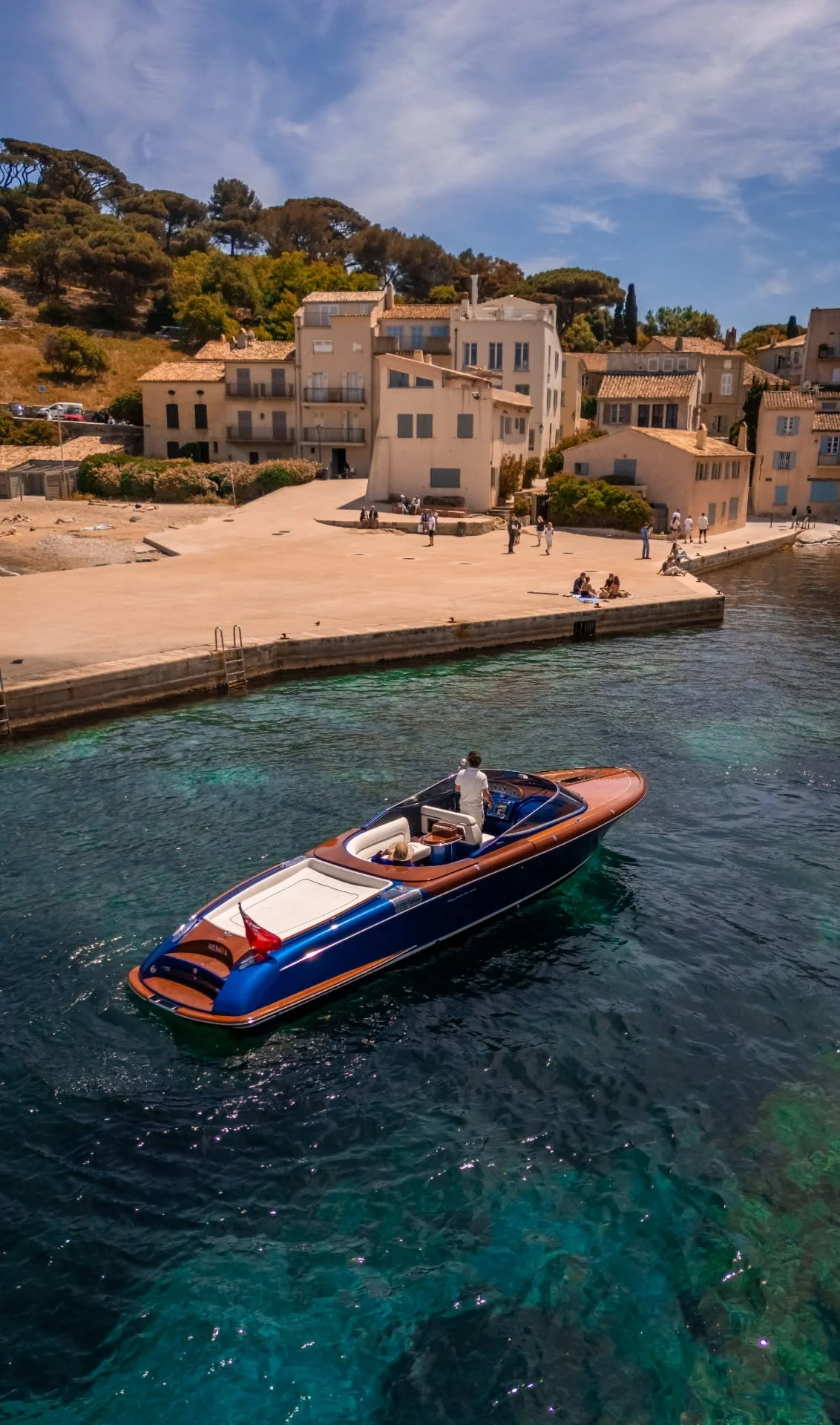 A speedboat with a wooden finish and dark blue hull floating on clear turquoise water near a Mediterranean-style village with white buildings and a stone pier.