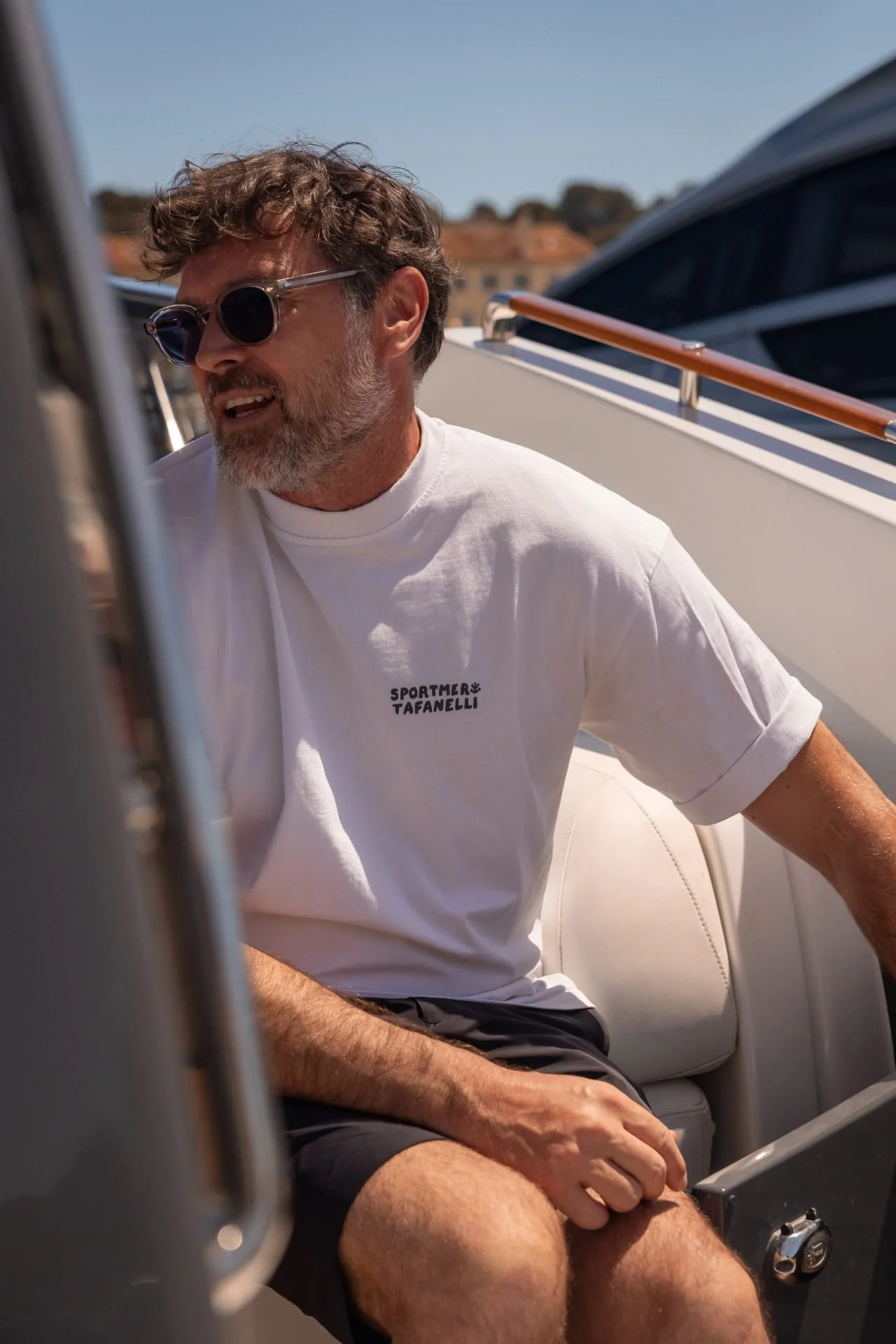 A man with curly hair and sunglasses sitting on a boat with a white t-shirt and black shorts, with buildings and a blue sky in the background.