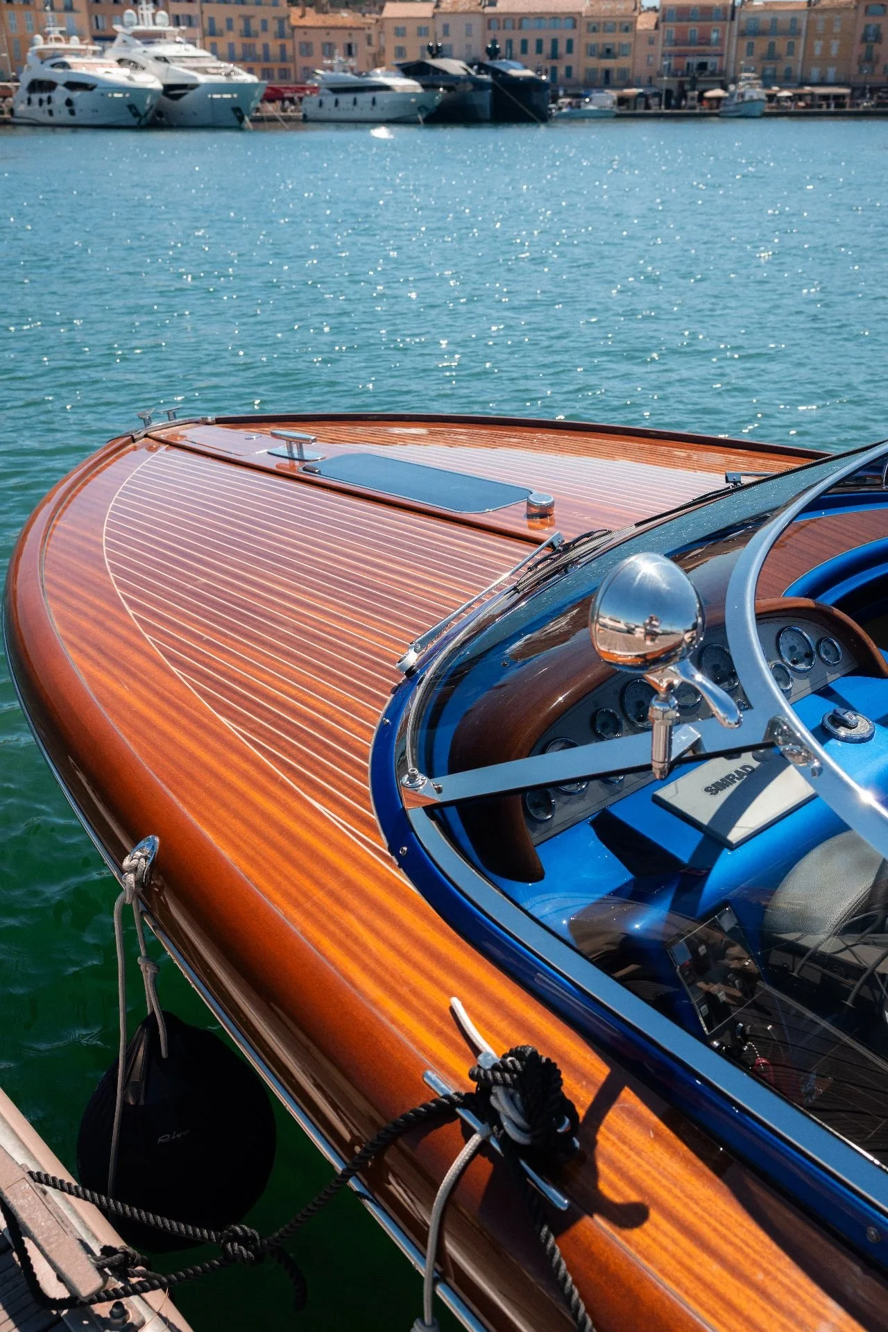 A close-up of a wooden speedboat's bow and cockpit on calm water, with a marina and buildings in the background.