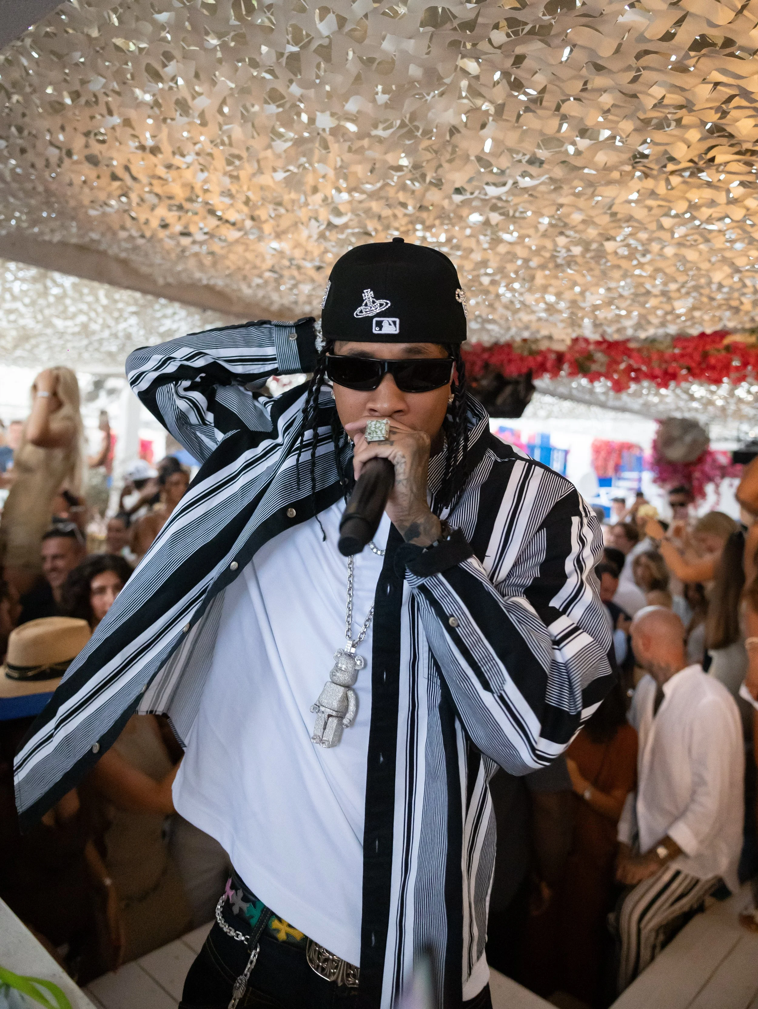 A man wearing a black baseball cap with a world logo, black sunglasses, a black and white striped jacket, and a large silver chain necklace is holding a microphone. He is in a crowded indoor space with a camouflage net ceiling, and other people are v