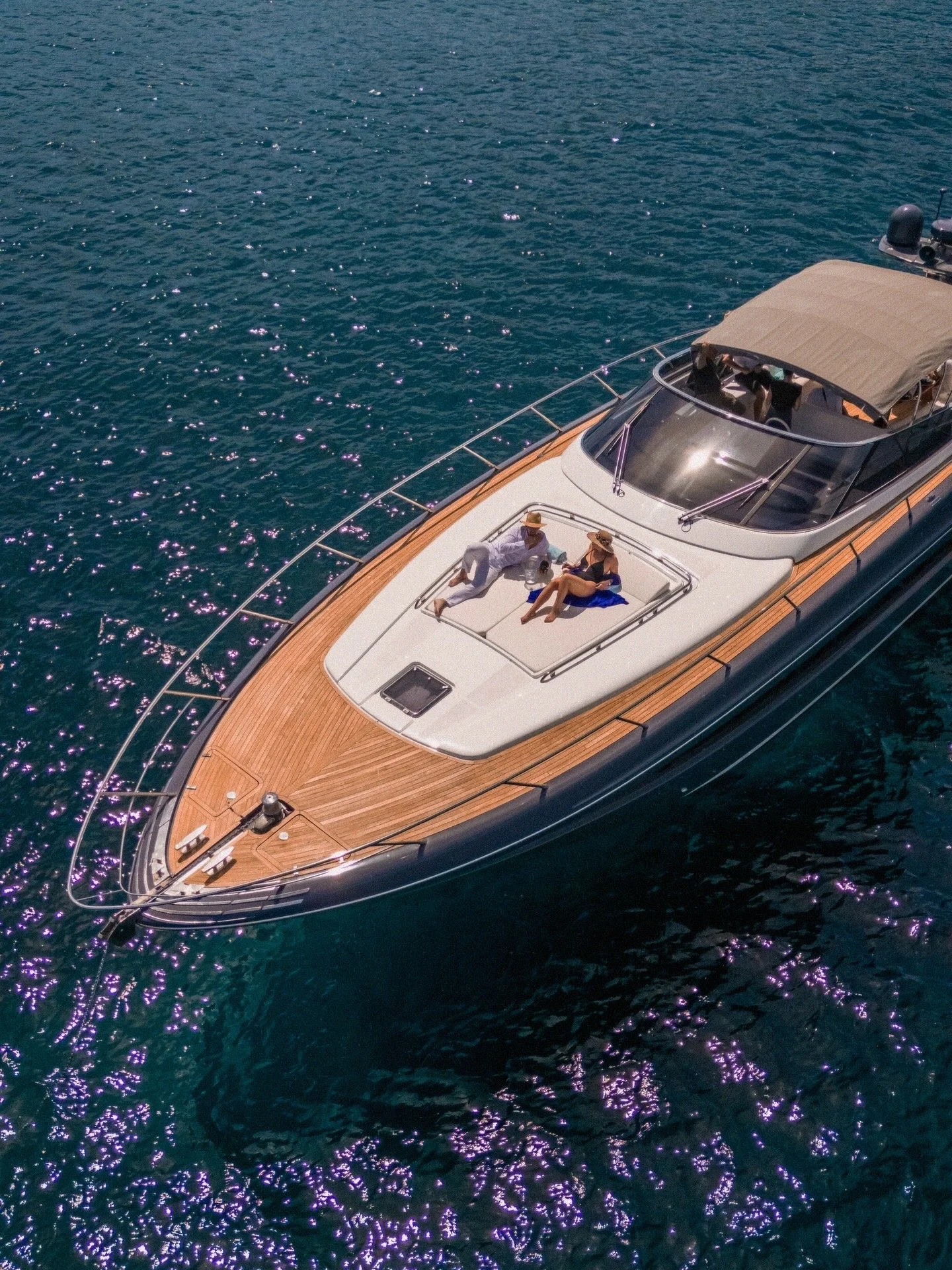 A yacht with a wooden deck and a gray canopy, floating on deep blue water with sunlight reflections, and two people relaxing on the deck.