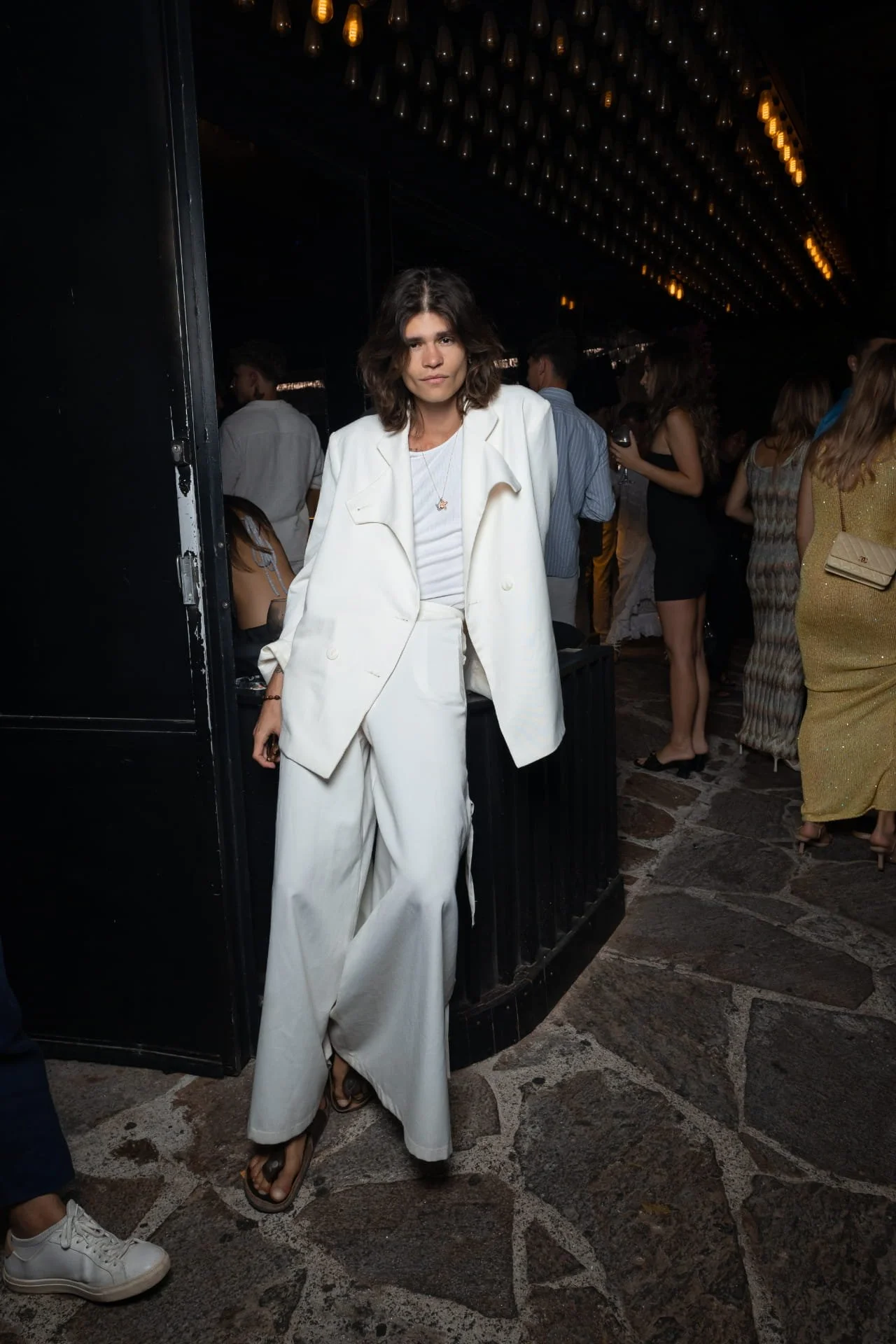 Woman with wavy brown hair in a white suit and white top, standing indoors at a social event, surrounded by people in stylish outfits, with warm lighting overhead.