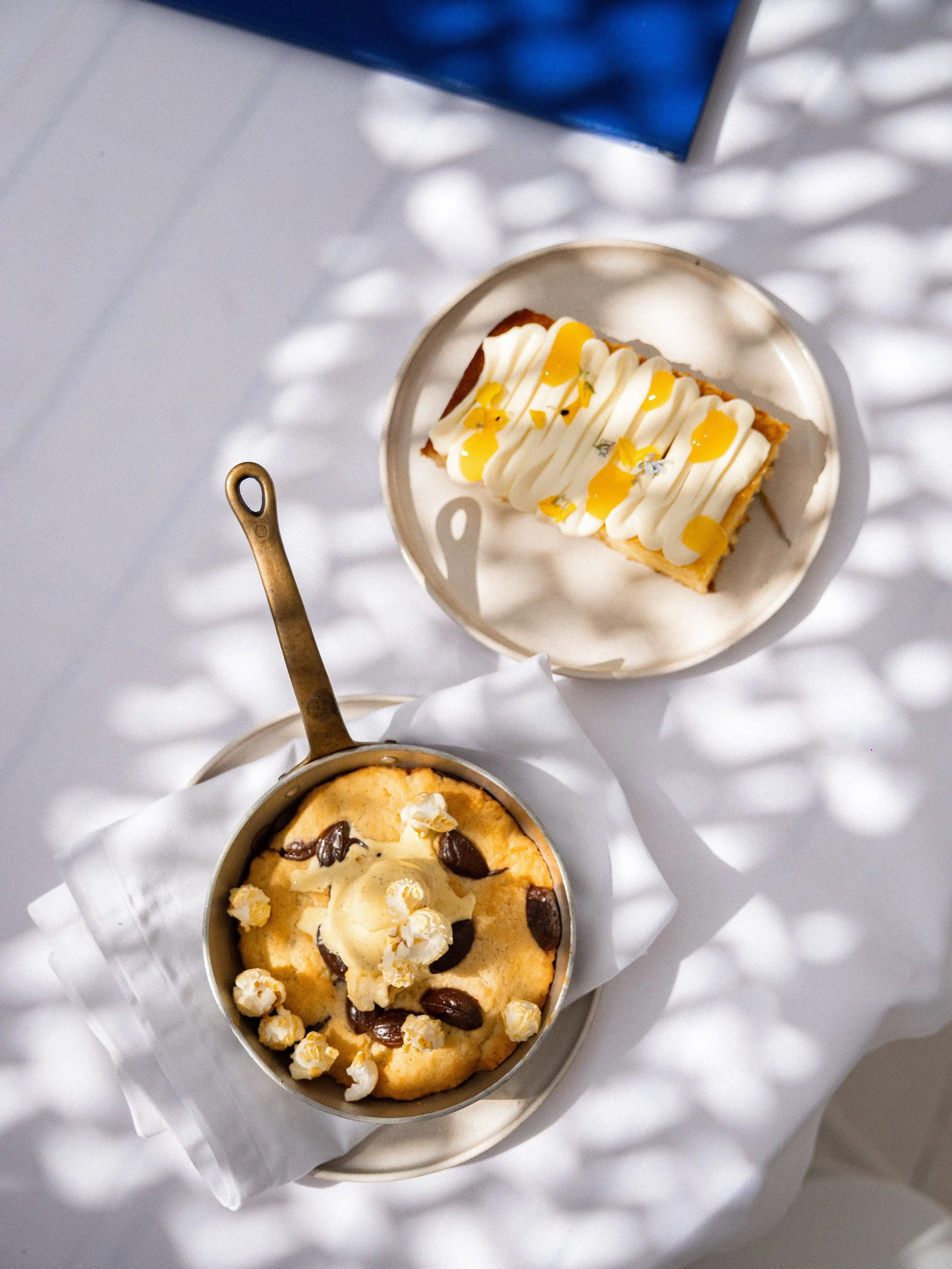 Two ice cream cups with toppings, one has popcorn and chocolate chips, the other has vanilla ice cream with popcorn, chocolate chips, and caramel drizzle, on a white surface with shadows.
