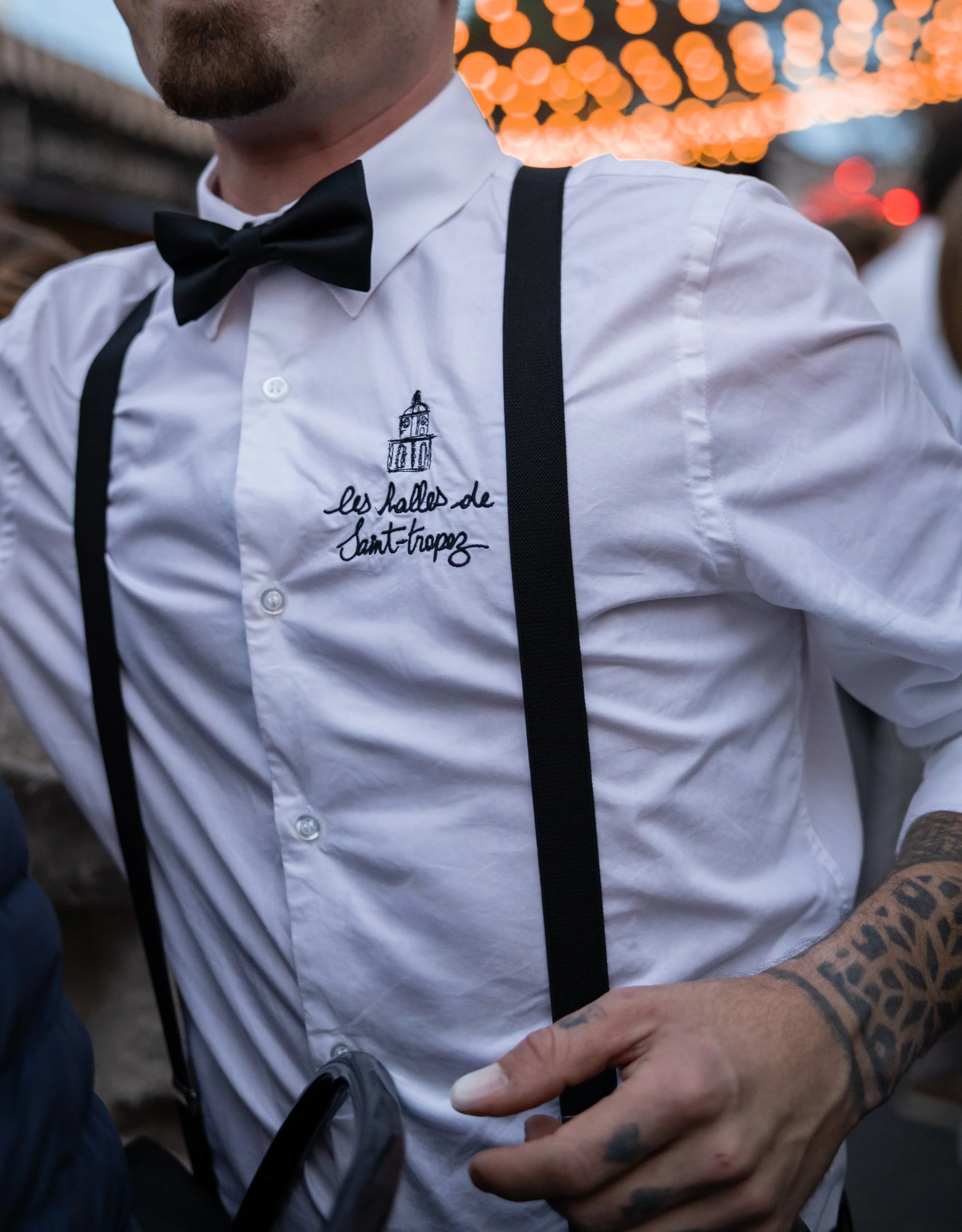 A close-up of a man wearing a white shirt with black suspenders, a black bow tie, and embroidered text and a drawing on the shirt. The man has a tattooed arm and is holding hands with another person. The background features out-of-focus orange string