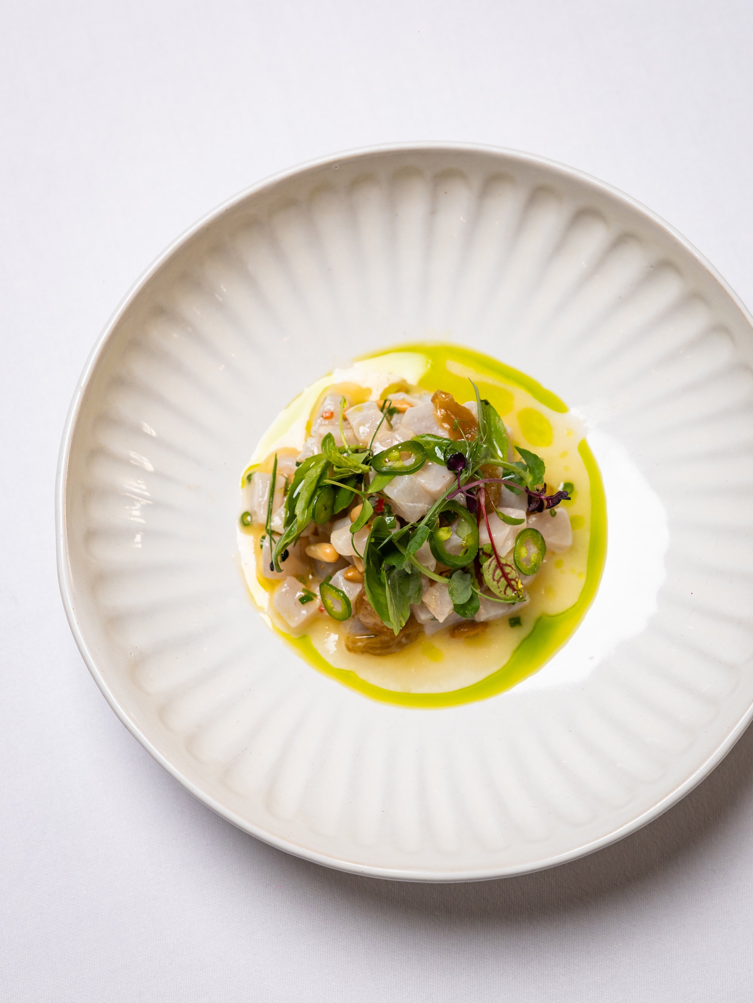 A white, round bowl with a textured interior rim containing a dish of diced seafood, garnished with microgreens and drizzled with yellow oil.