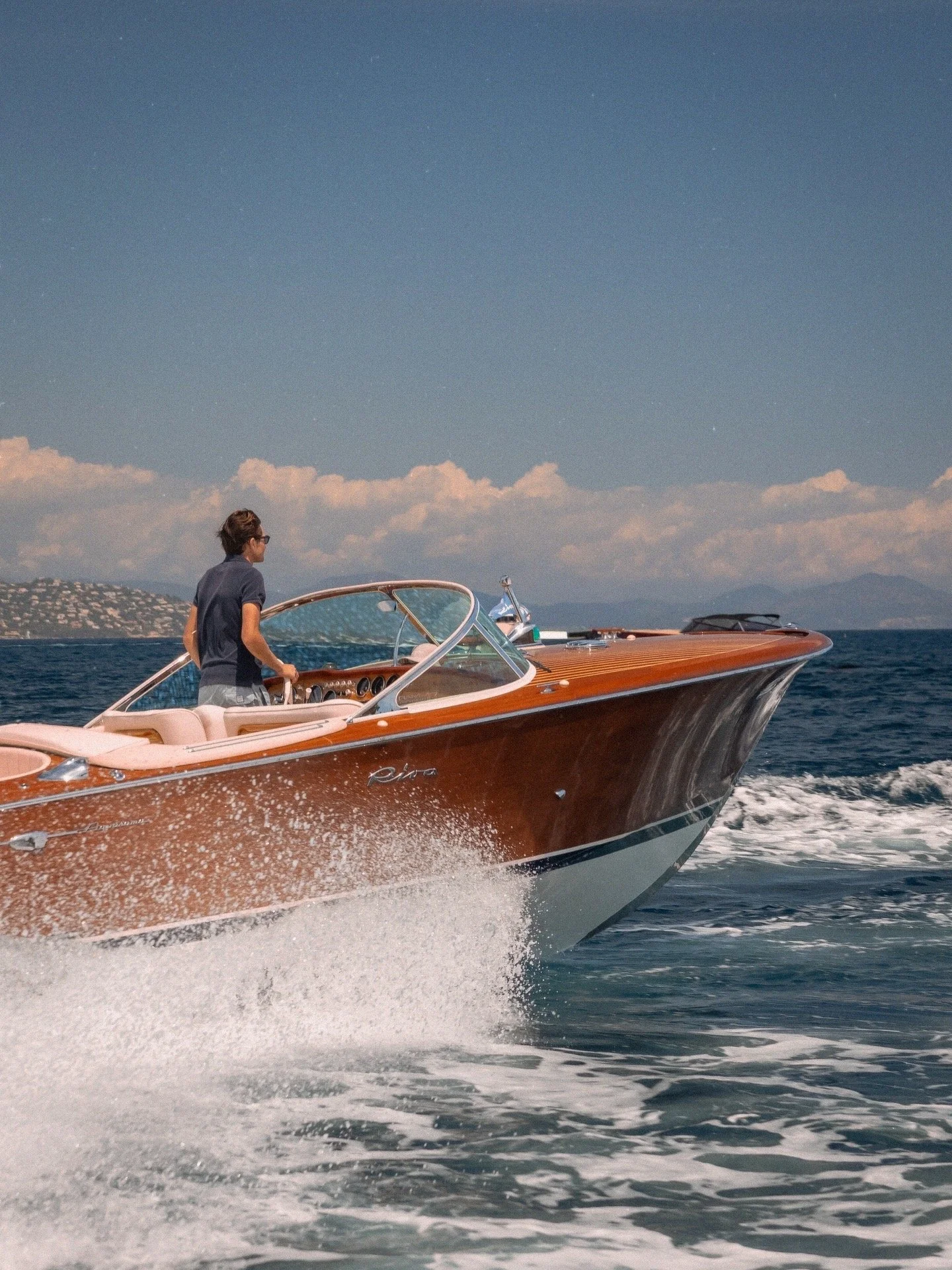 A man wearing glasses and a dark shirt driving a wooden speedboat on the water, with a city and mountains in the background under a partly cloudy sky.