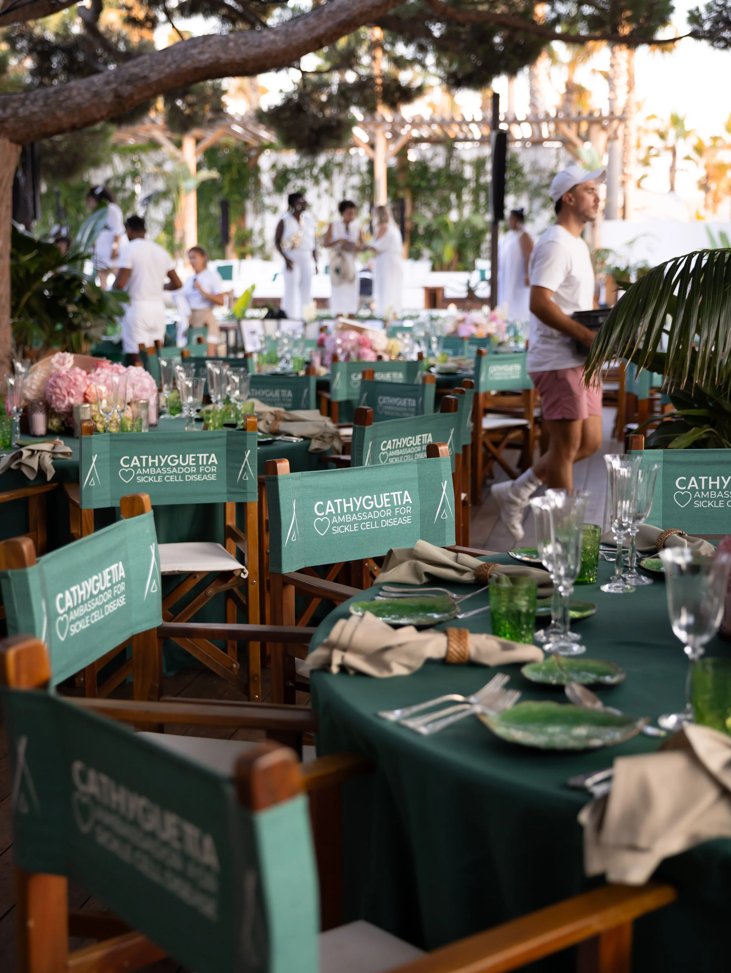Outdoor event with tables decorated with pink flowers and green tablecloths, chairs with signs supporting Sickle Cell Disease awareness, and staff members setting up or attending to the preparations, under trees during sunset.