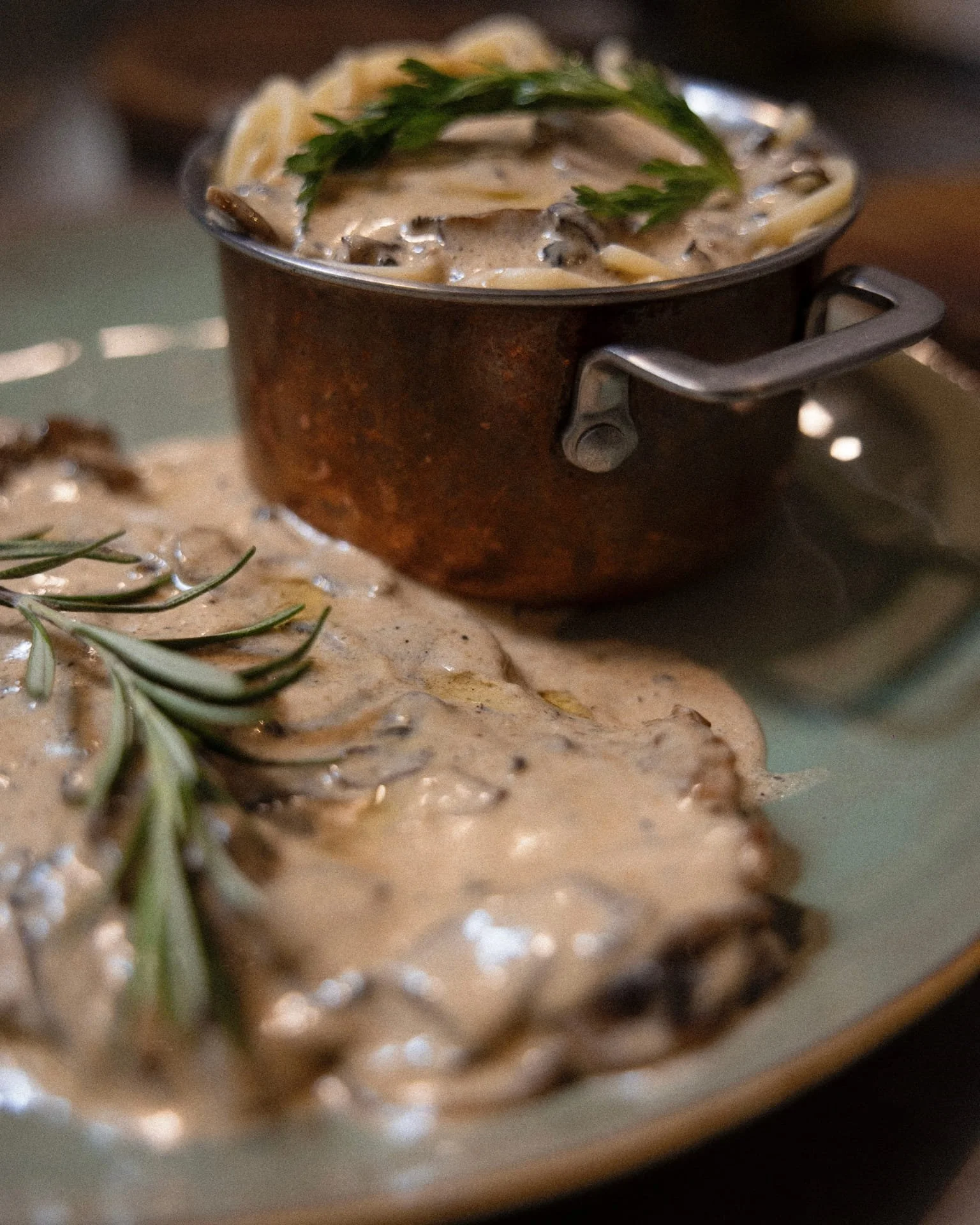 A small metal cup filled with creamy mushroom soup garnished with a sprig of fresh parsley, placed on a patterned surface with a sprig of rosemary nearby.