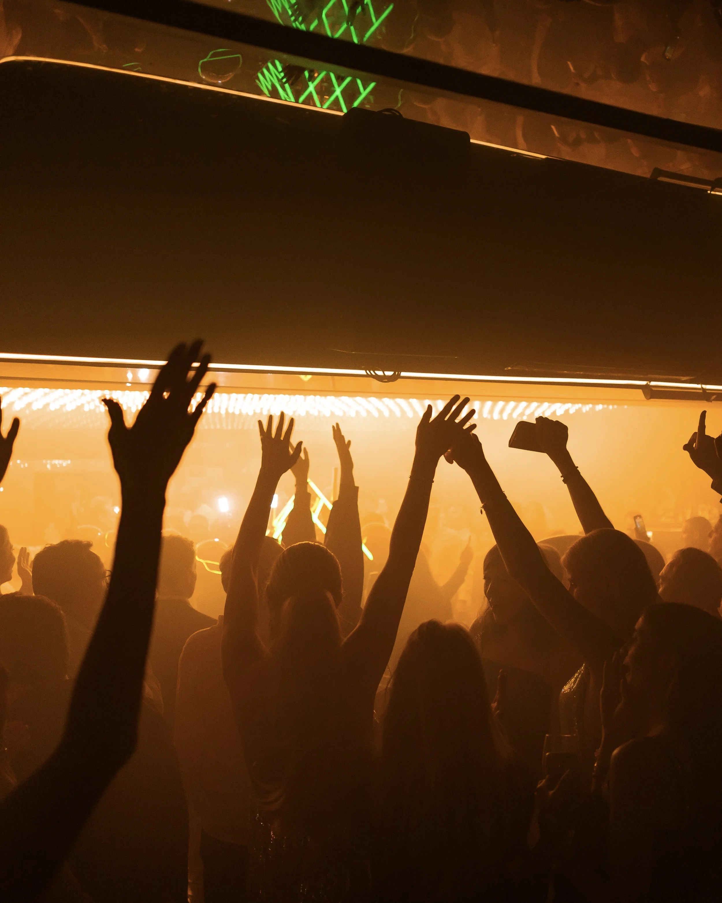Crowd of people raising their hands at a concert or music event with a stage illuminated in orange light and visible green and yellow neon lights in the background.