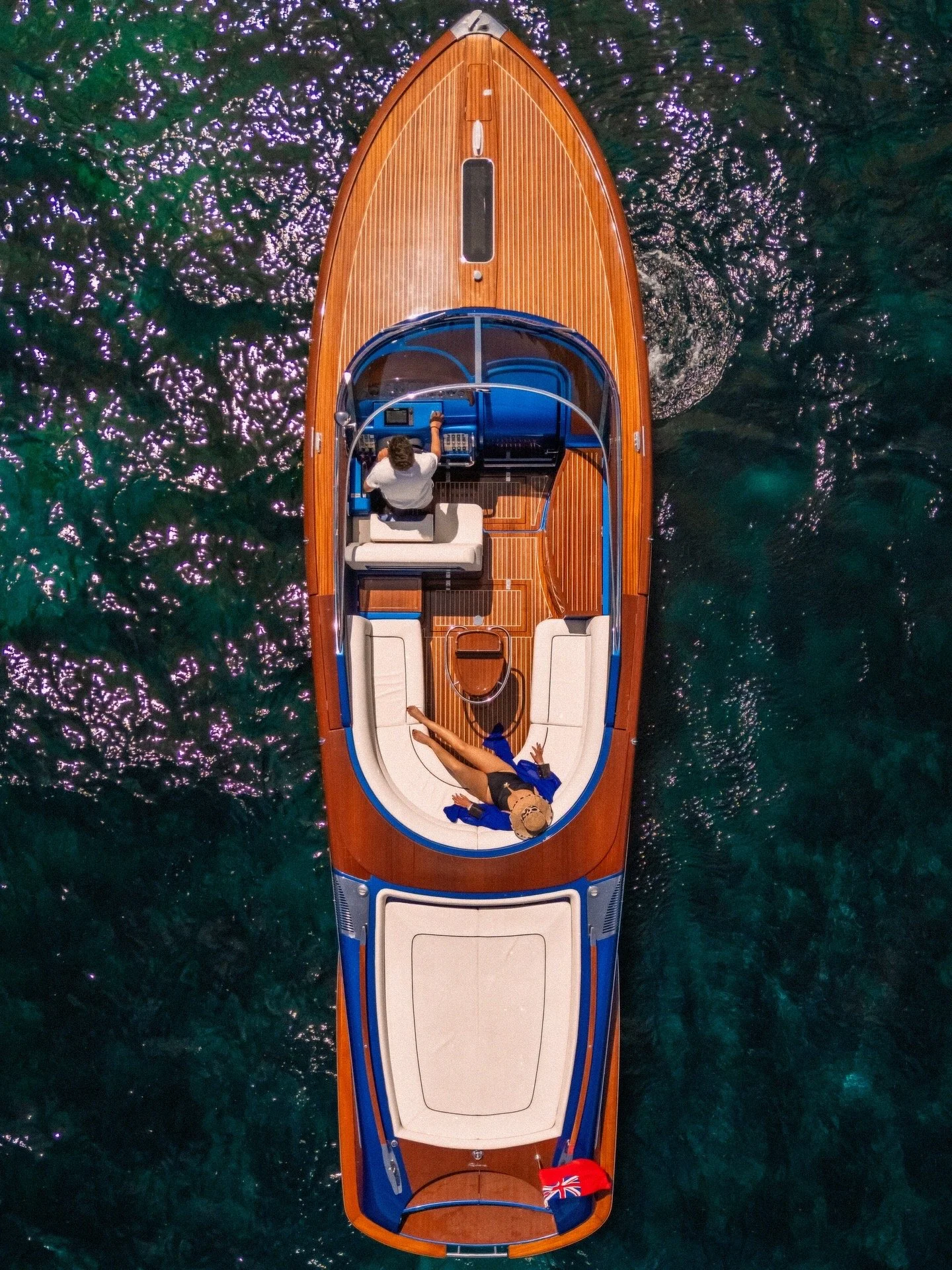 Top-down view of a wooden yacht on clear water, with a person lying on a white lounge at the front and a person operating the boat's controls at the rear, with a small British flag at the back.