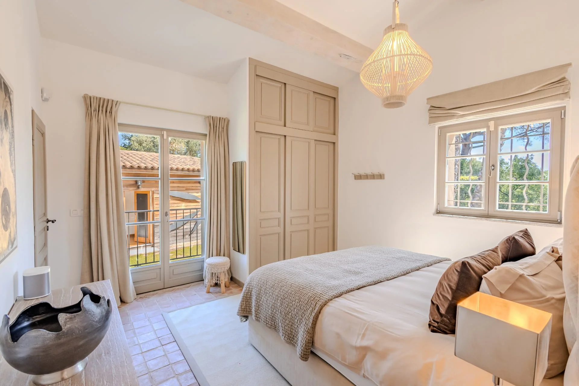 A cozy bedroom with white walls, a large bed with beige and brown pillows, a beige blanket, and a small nightstand with a lamp. There is a window with curtains and a view of trees outside, a built-in wardrobe, a ceiling light, and decorative items li