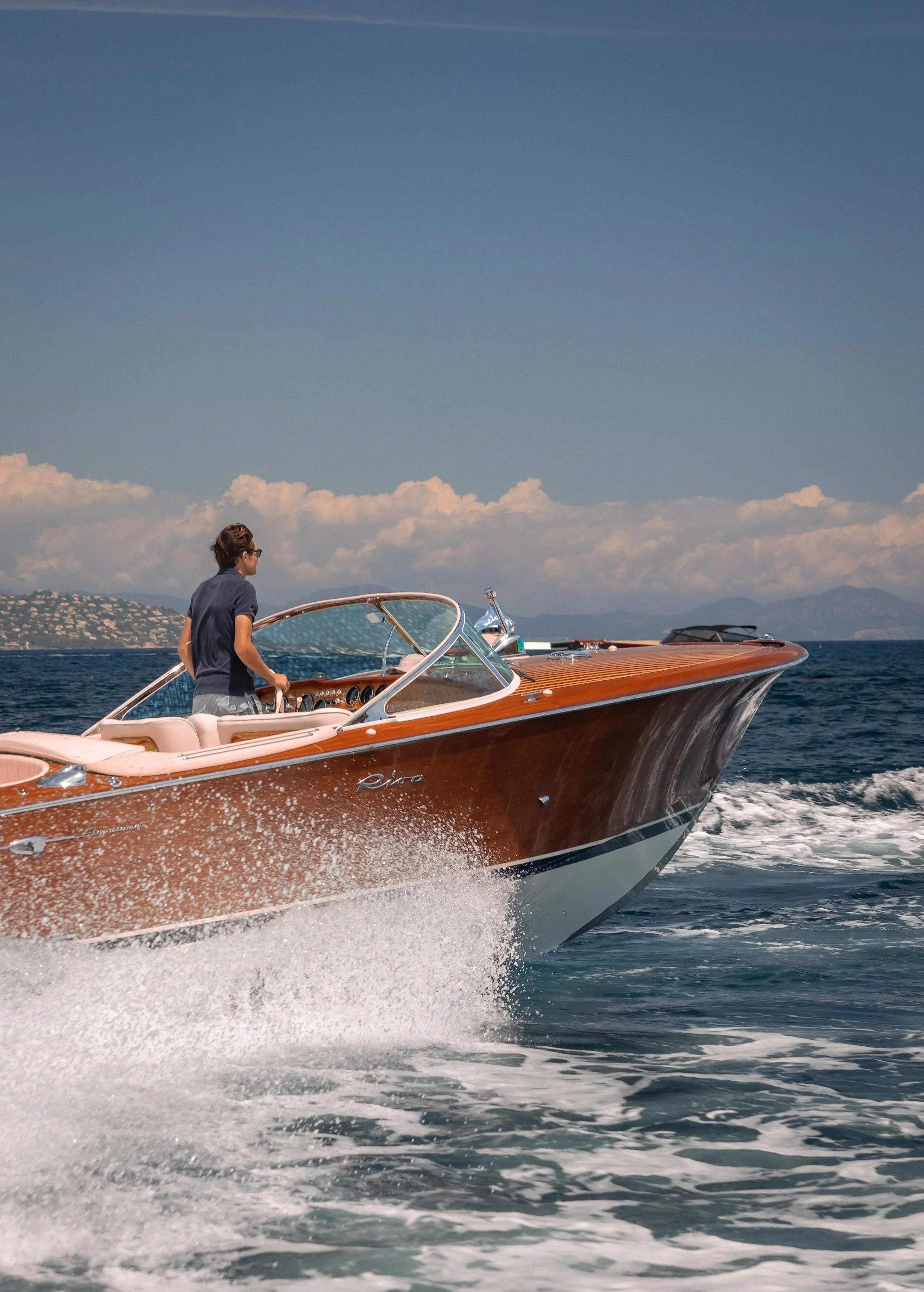 A man wearing a dark shirt and shorts is driving a wooden speedboat that is cutting through water. The scene is set against a backdrop of distant hills, mountains, and a sky filled with clouds.