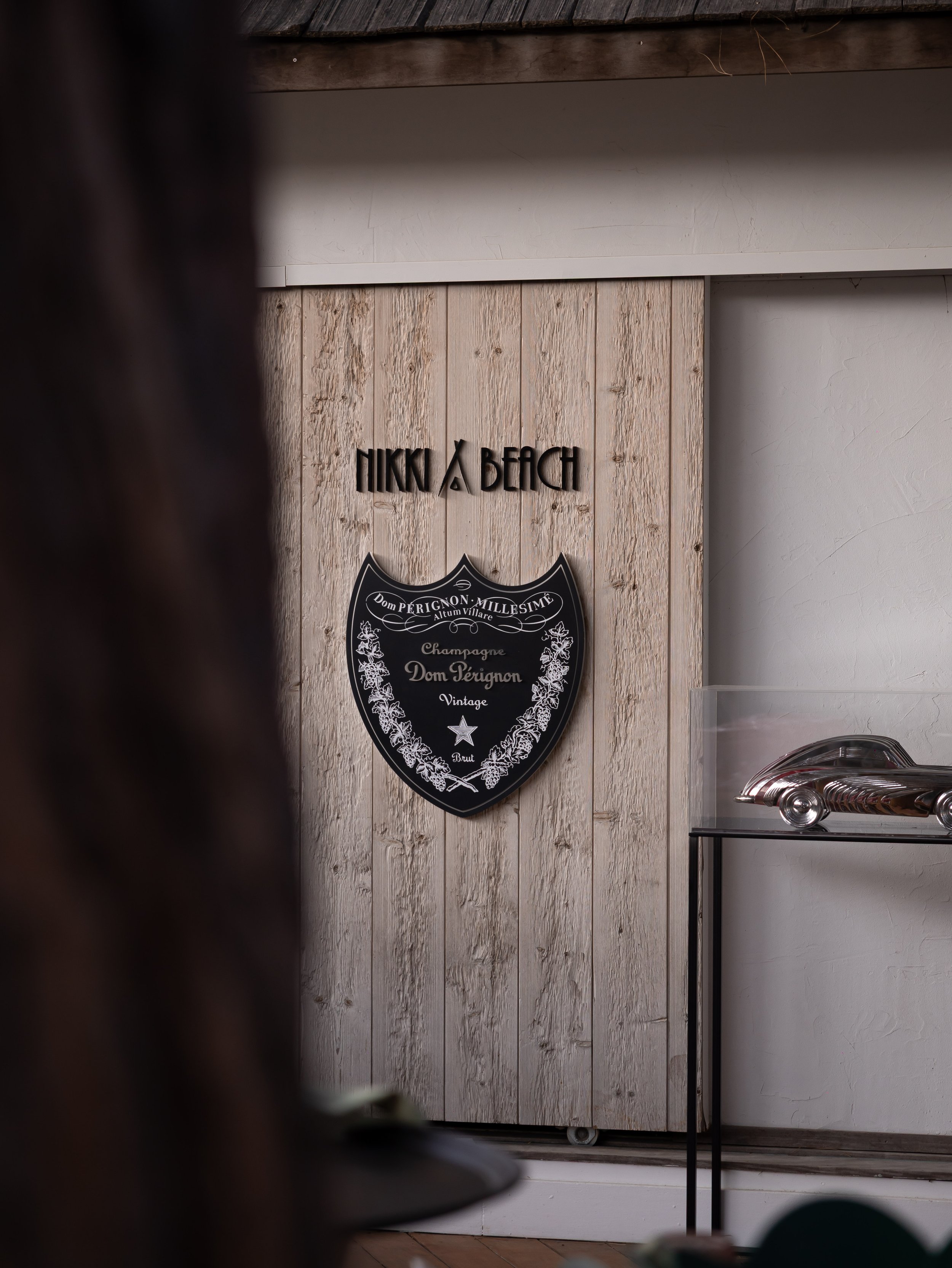 Photo taken from a low angle showing a wall with a decorative sign that reads 'Nikki Beach' and a black shield-shaped plaque below it that says 'Dom Pérignon Millésimé Champagne Vintage.' Part of a metal stand and a glass display case are visible in 