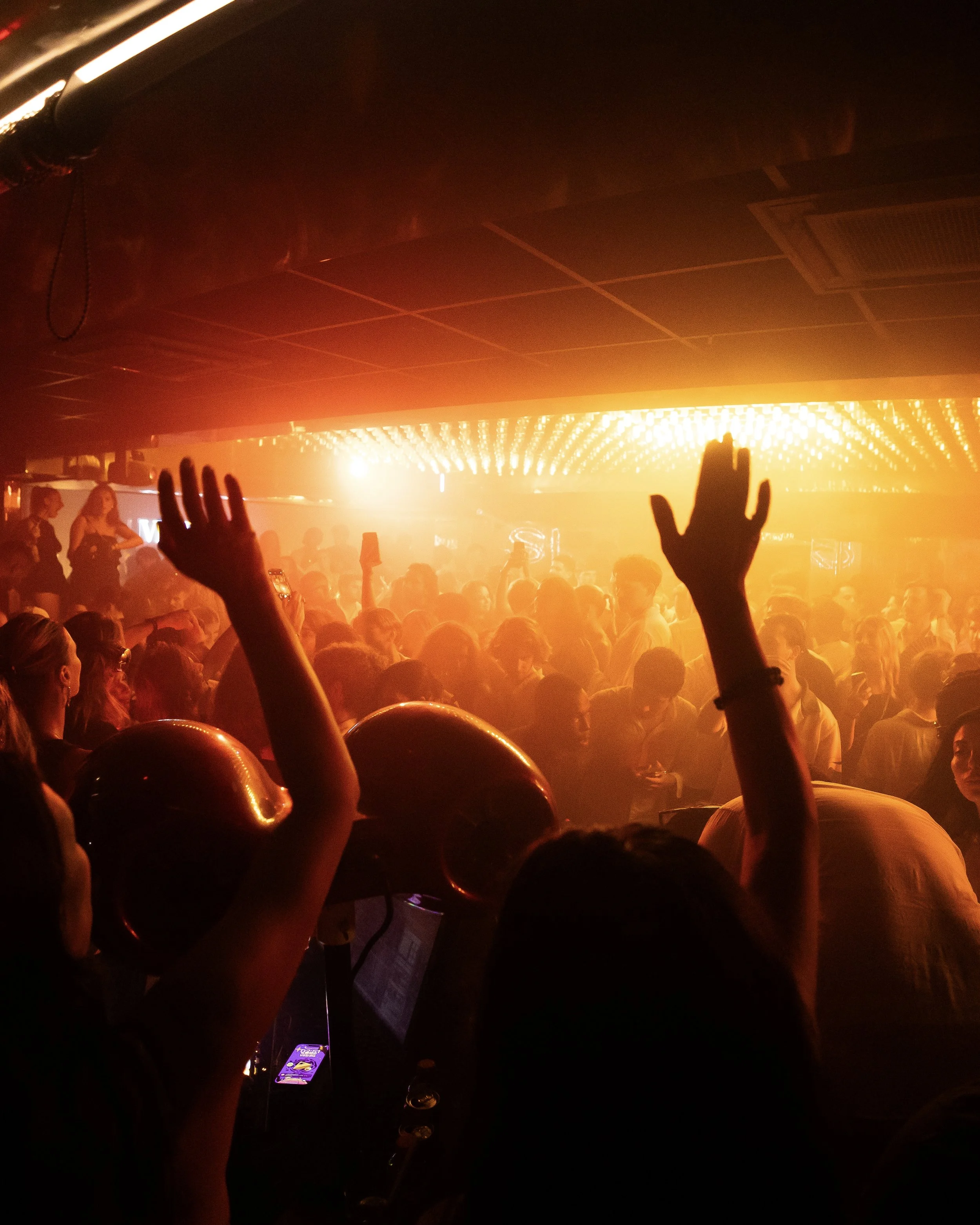 Crowd of people at concert with raised hands, dark indoor setting, illuminated ceiling lights, and a bright stage in the background.
