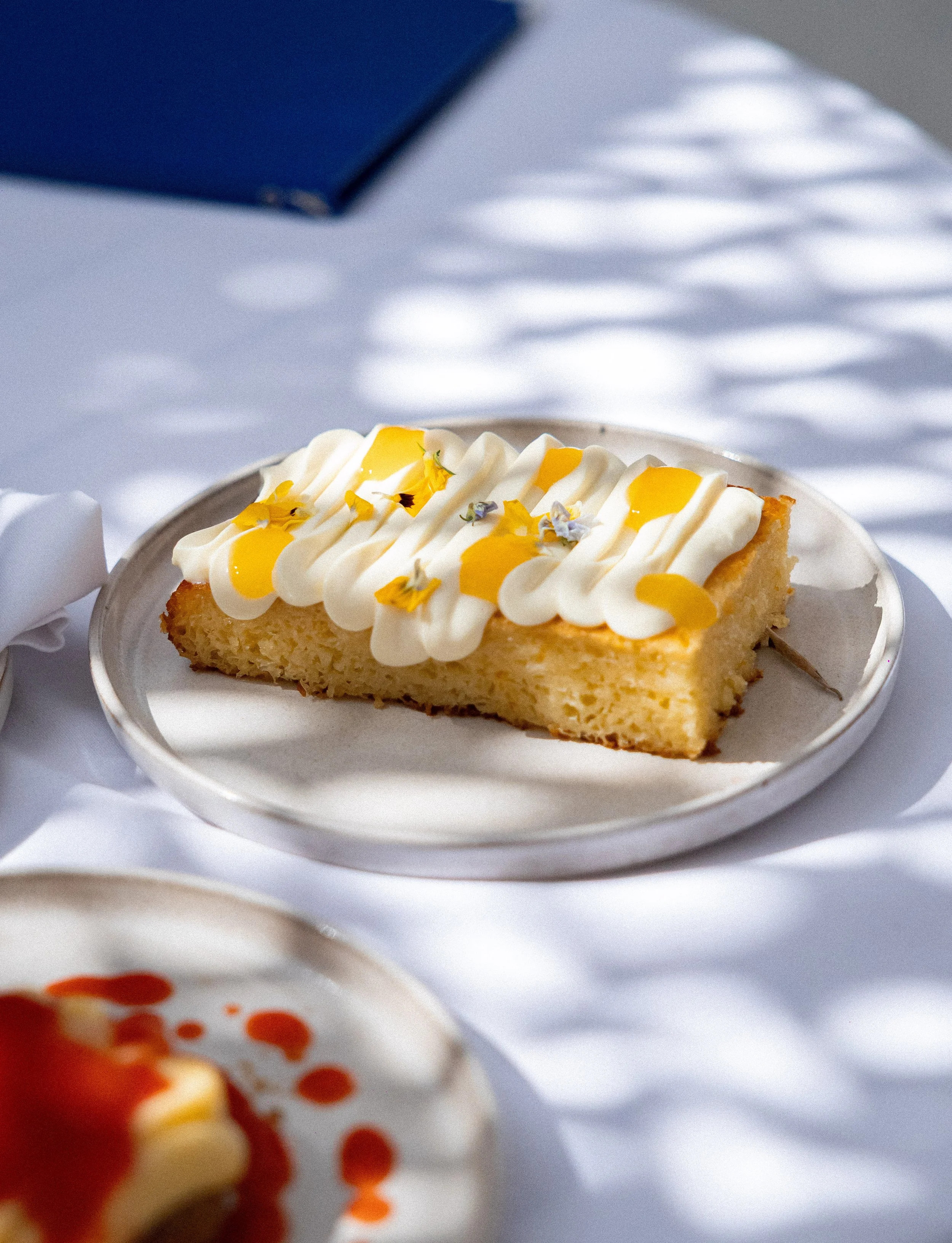 A slice of cheesecake with white cream topping, yellow sauce, and edible flowers, served on a white plate. In the background, part of another dessert with red sauce and white cream.