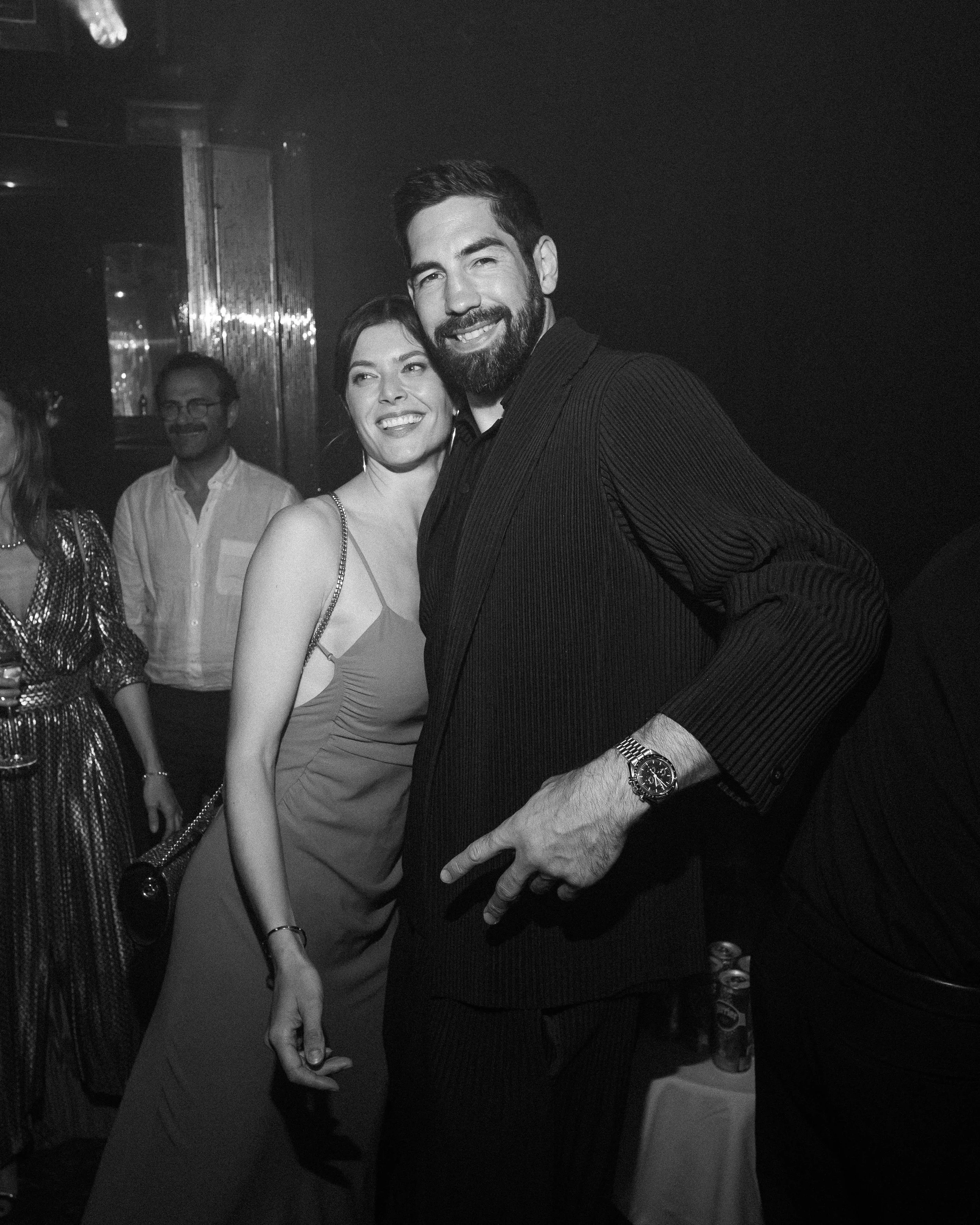 A black and white photograph of a group of people at a social gathering or party, with two individuals in the foreground smiling and standing close together, and others in the background.
