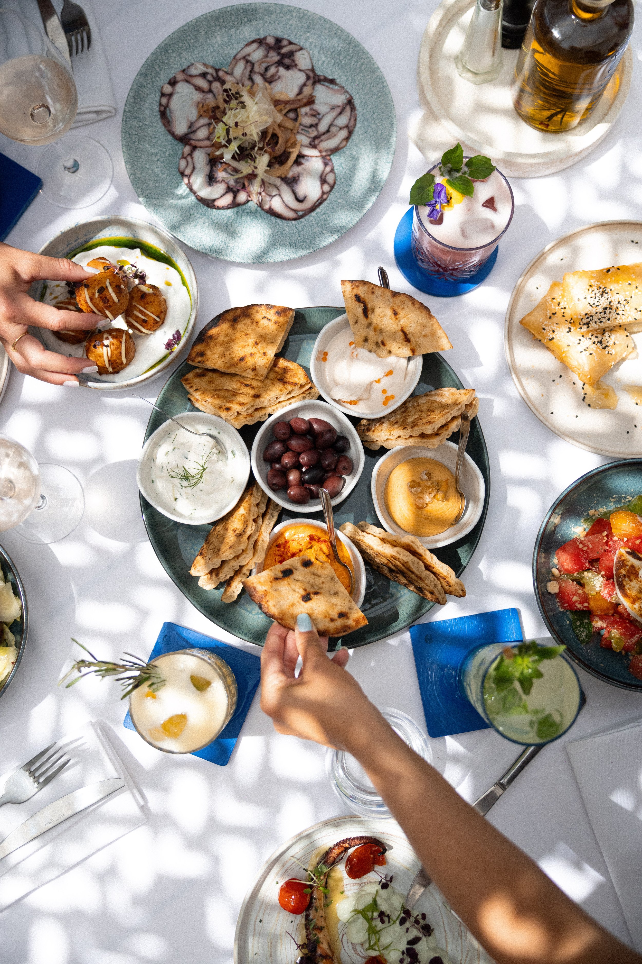 A top-down view of a table filled with various dishes of food and drinks. There are plates of grilled flatbreads, a bowl of olives, a plate with sliced cured meat, a small bowl of creamy dip, and bowls of cheese and salads. Hands are reaching for foo