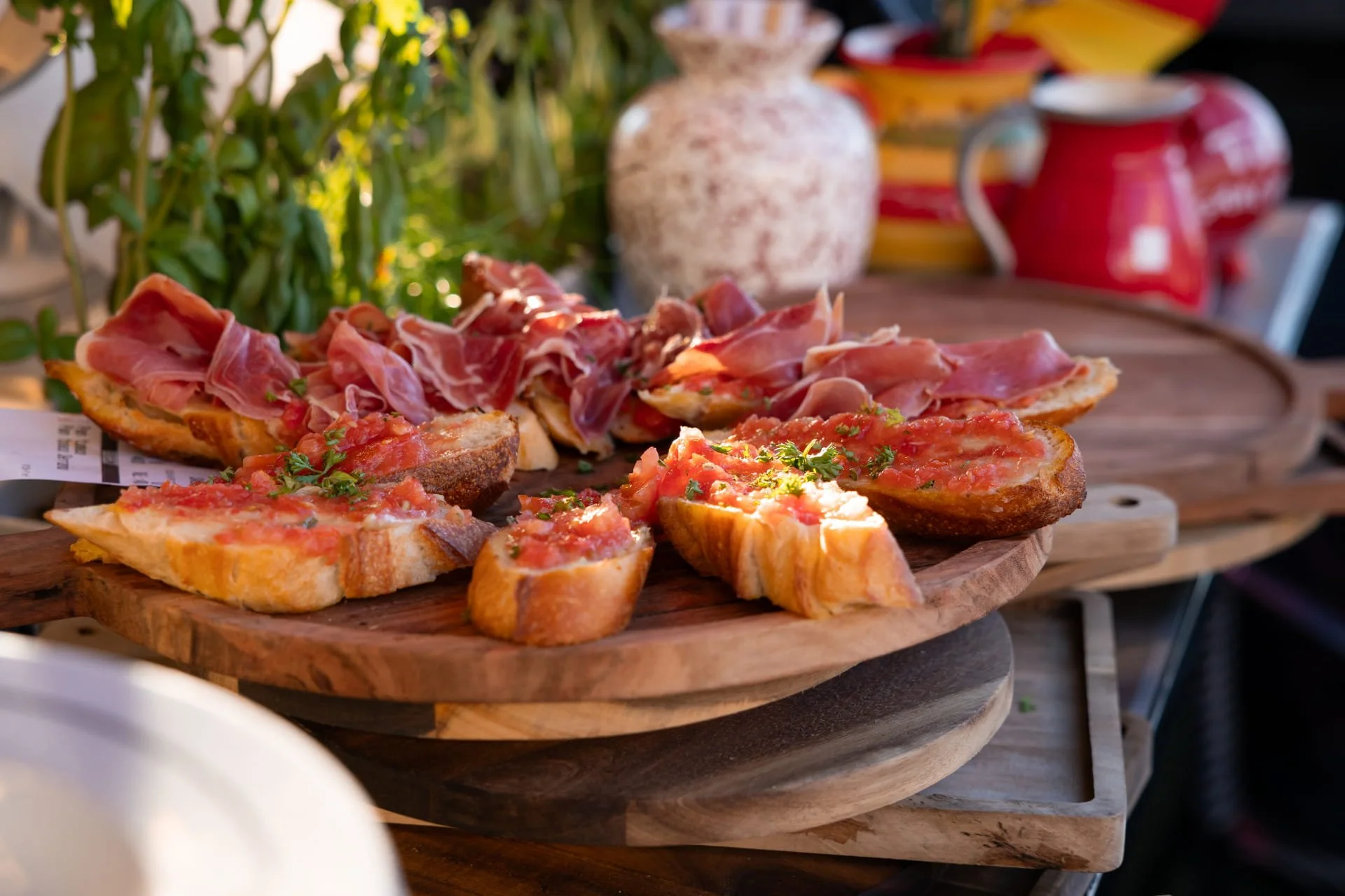 Wooden serving platter with bruschetta topped with tomato and herbs, and slices of prosciutto on toasted baguette slices, with blurred pottery and plants in the background.