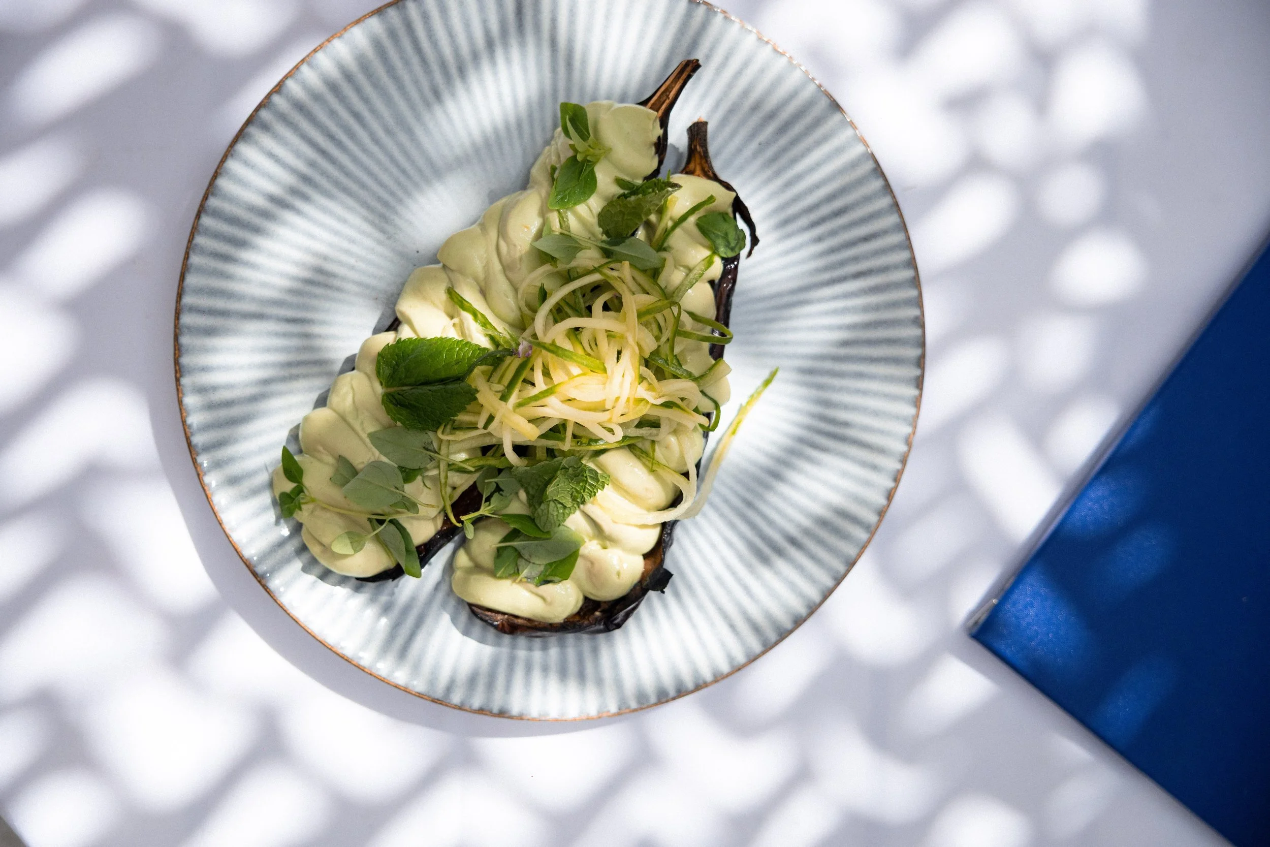 A dish featuring roasted eggplant topped with creamy avocado sauce, shredded cheese, and fresh herbs, served on a decorative oval plate.