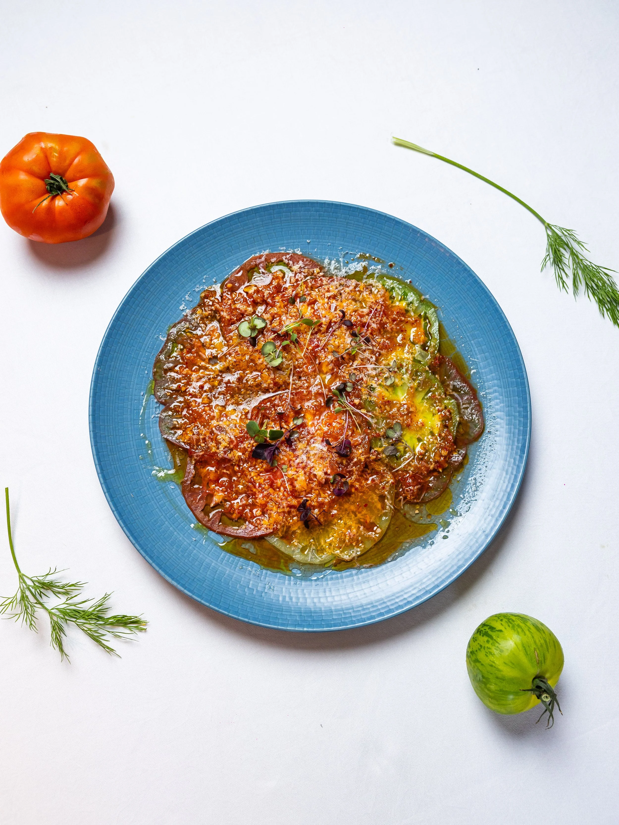A blue plate with a baked vegetable dish topped with microgreens, placed on a white surface. To the left of the plate is an orange heirloom tomato, to the right are two green tomatoes, and there are sprigs of dill on the left and right edges of the i