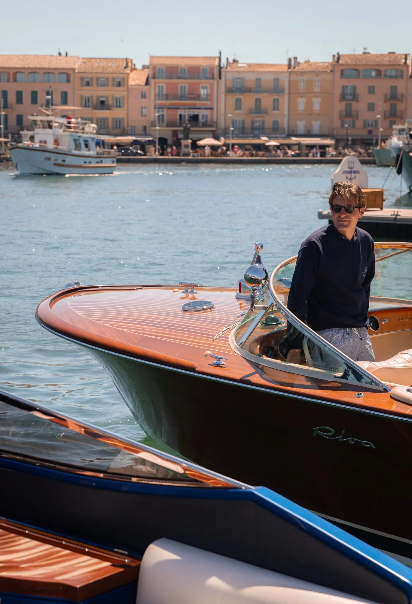 A man with sunglasses standing on a wooden boat in a harbor with colorful buildings and other boats in the background.
