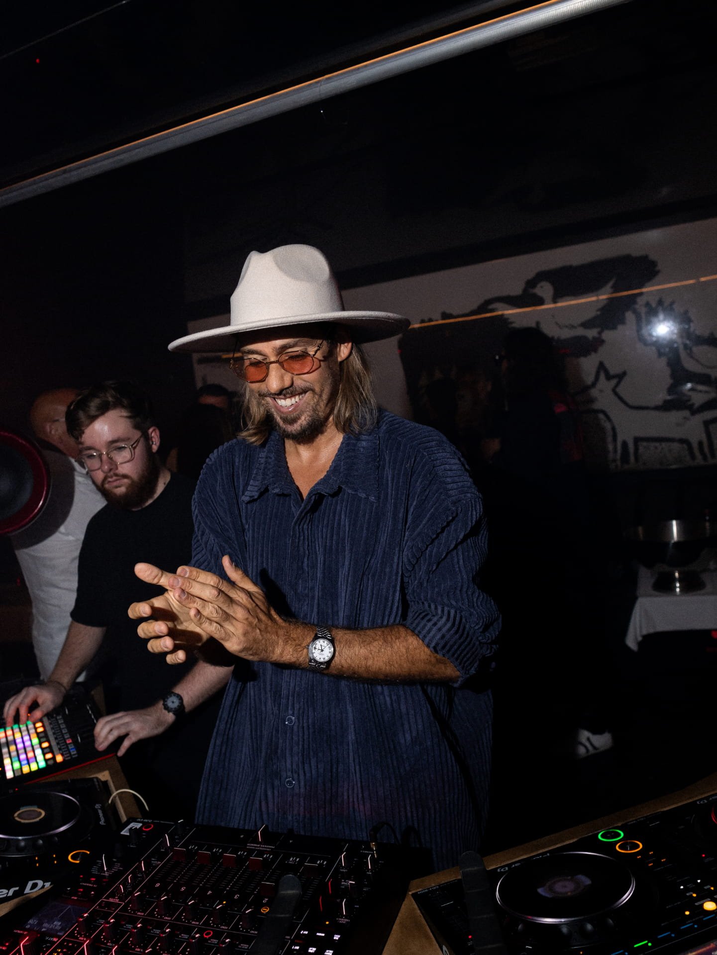 Man with long hair, glasses, and a white wide-brimmed hat smiling in a dimly lit room with DJ equipment, surrounded by other people.