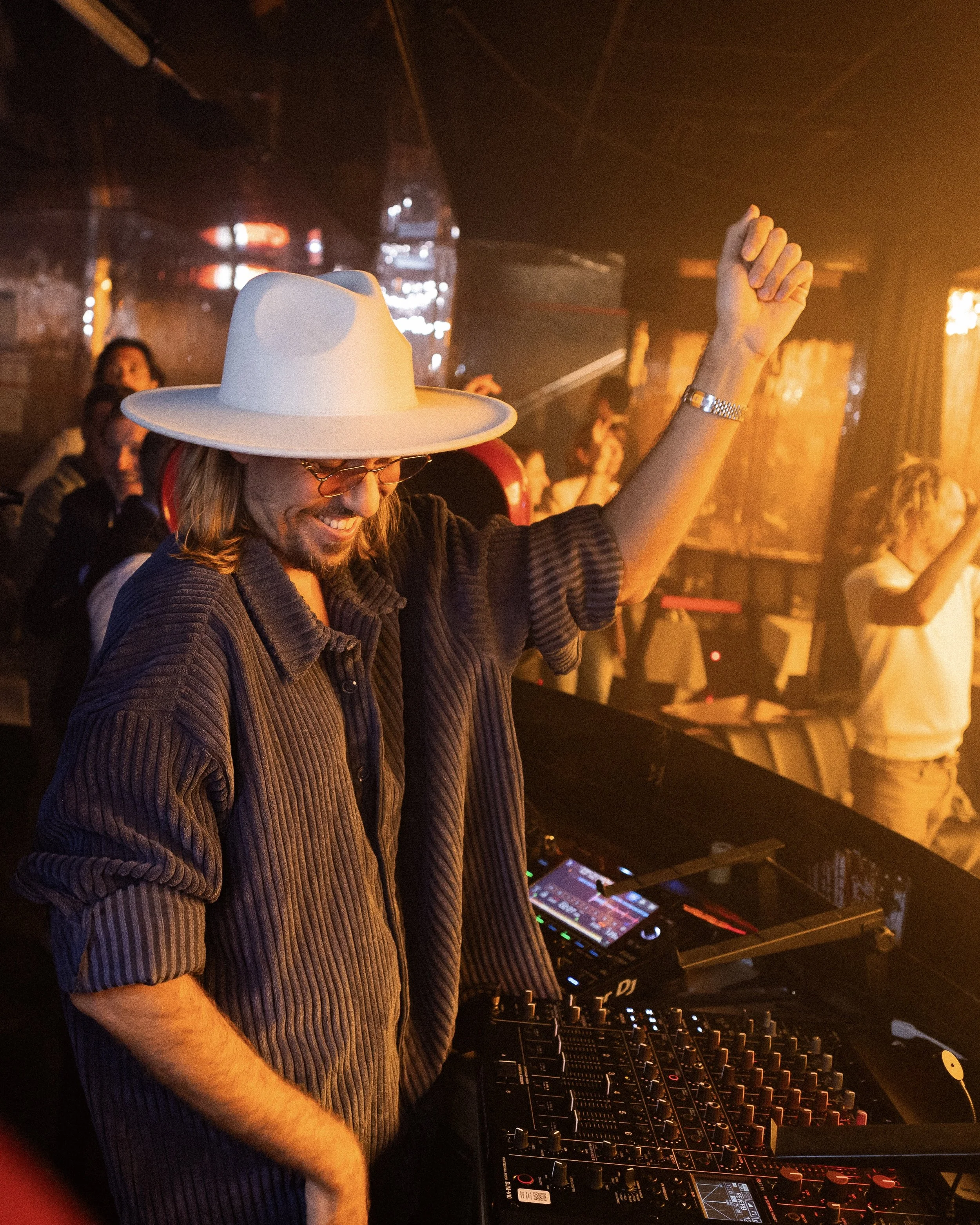 A DJ wearing a white hat and glasses, smiling, with a raised hand at a music event. The DJ is surrounded by DJ equipment, including turntables and a mixer, with warm lighting and a dark background.