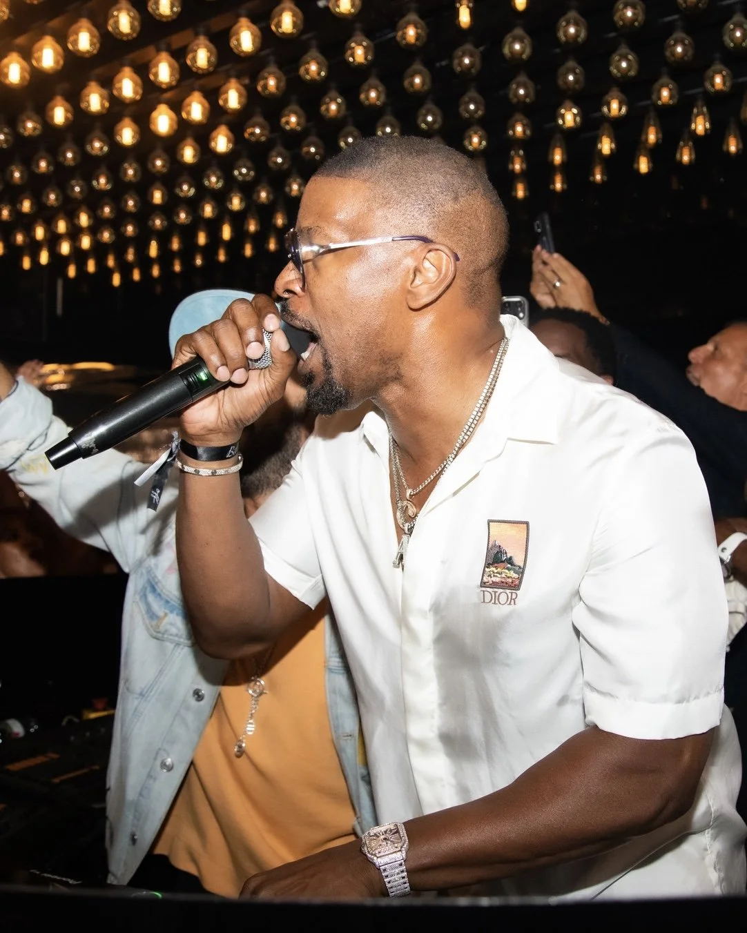 A man singing into a microphone at a party or event with multiple hanging lights in the background.