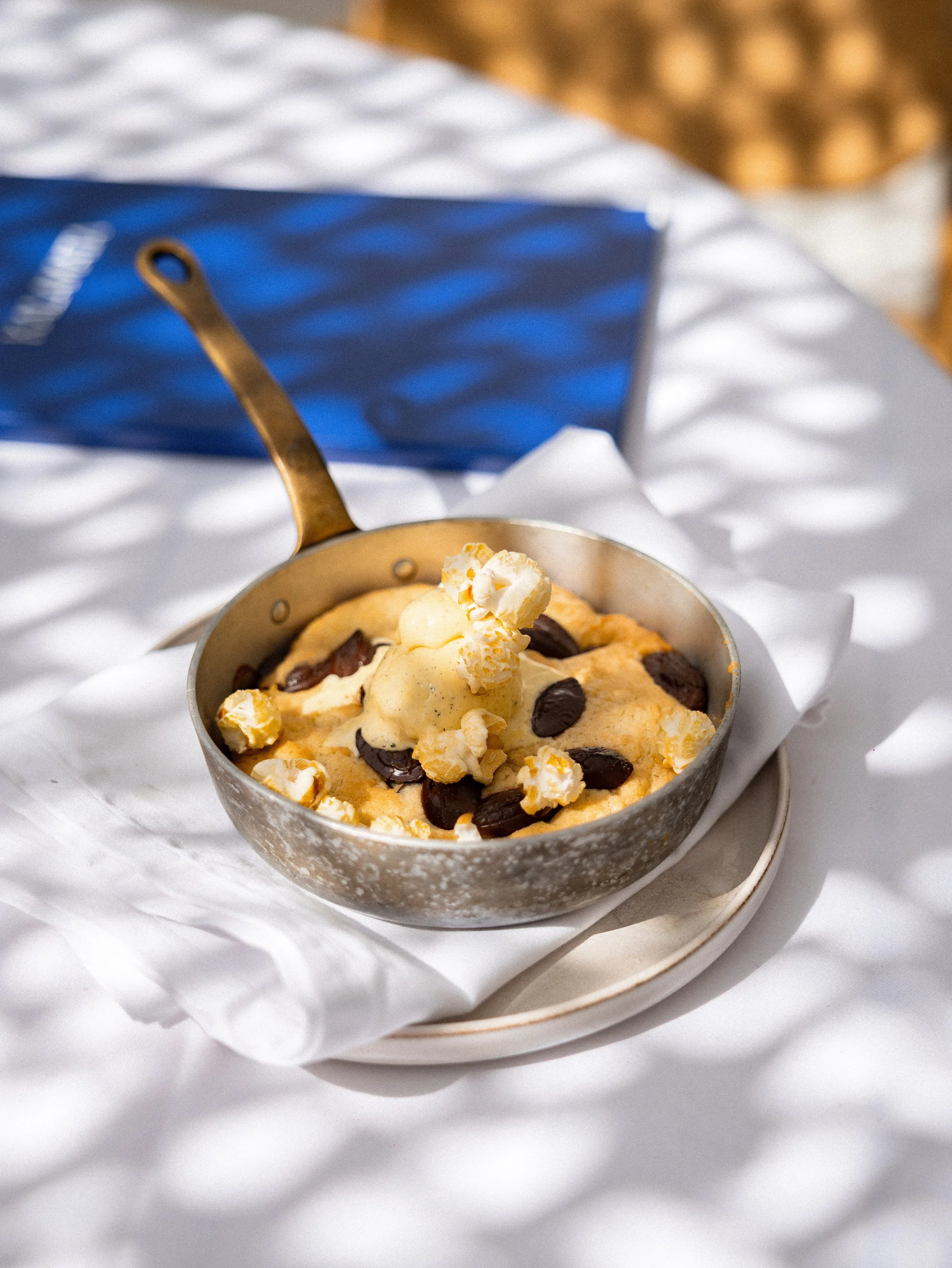 A bowl of cookie dough with chocolate chips and a dollop of whipped cream or butter on top, placed on a white plate with a handle, on a white cloth surface.