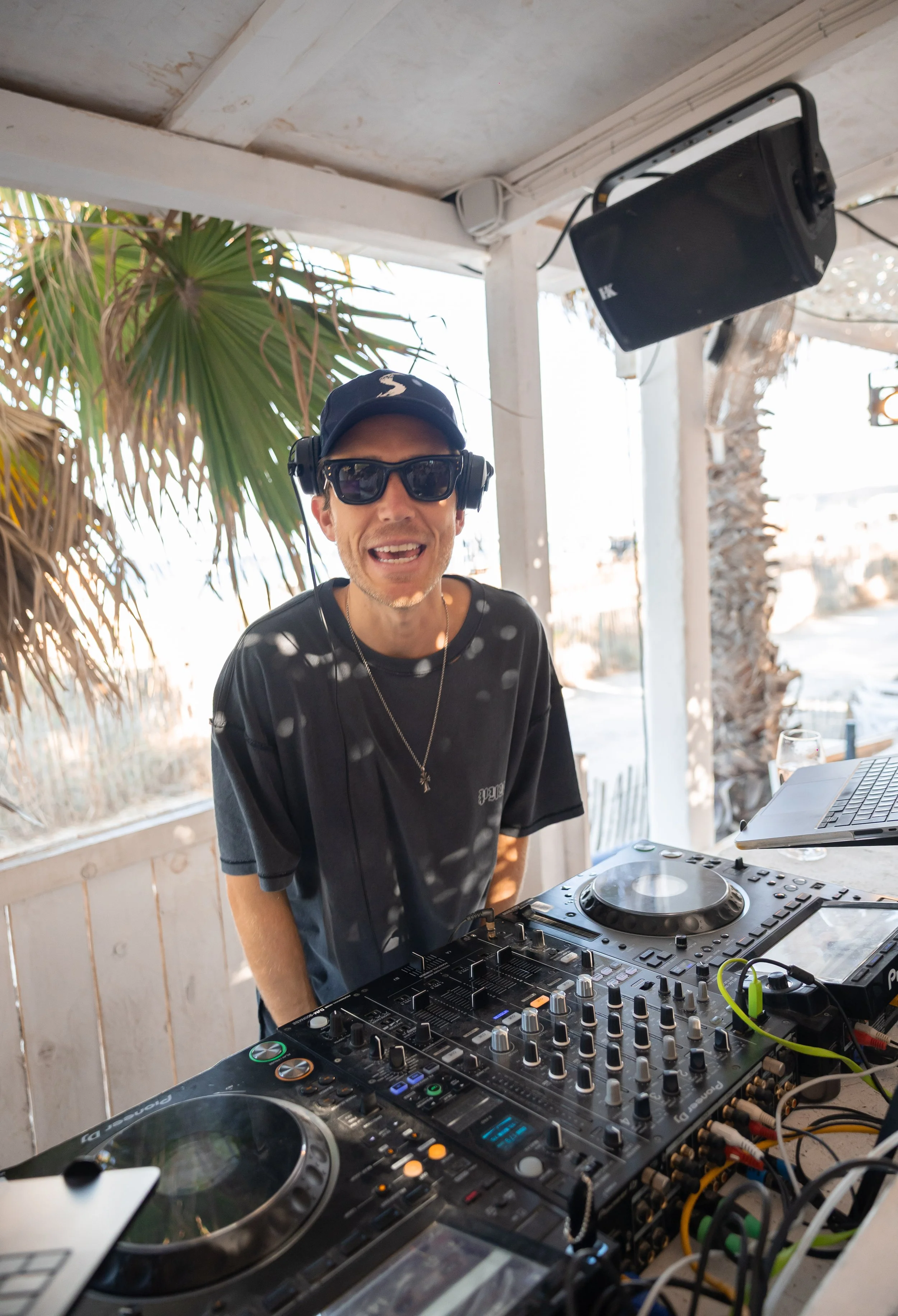 A DJ wearing sunglasses and headphones, smiling, standing behind a DJ mixer and turntables outdoors with palm trees and a bright sky in the background.