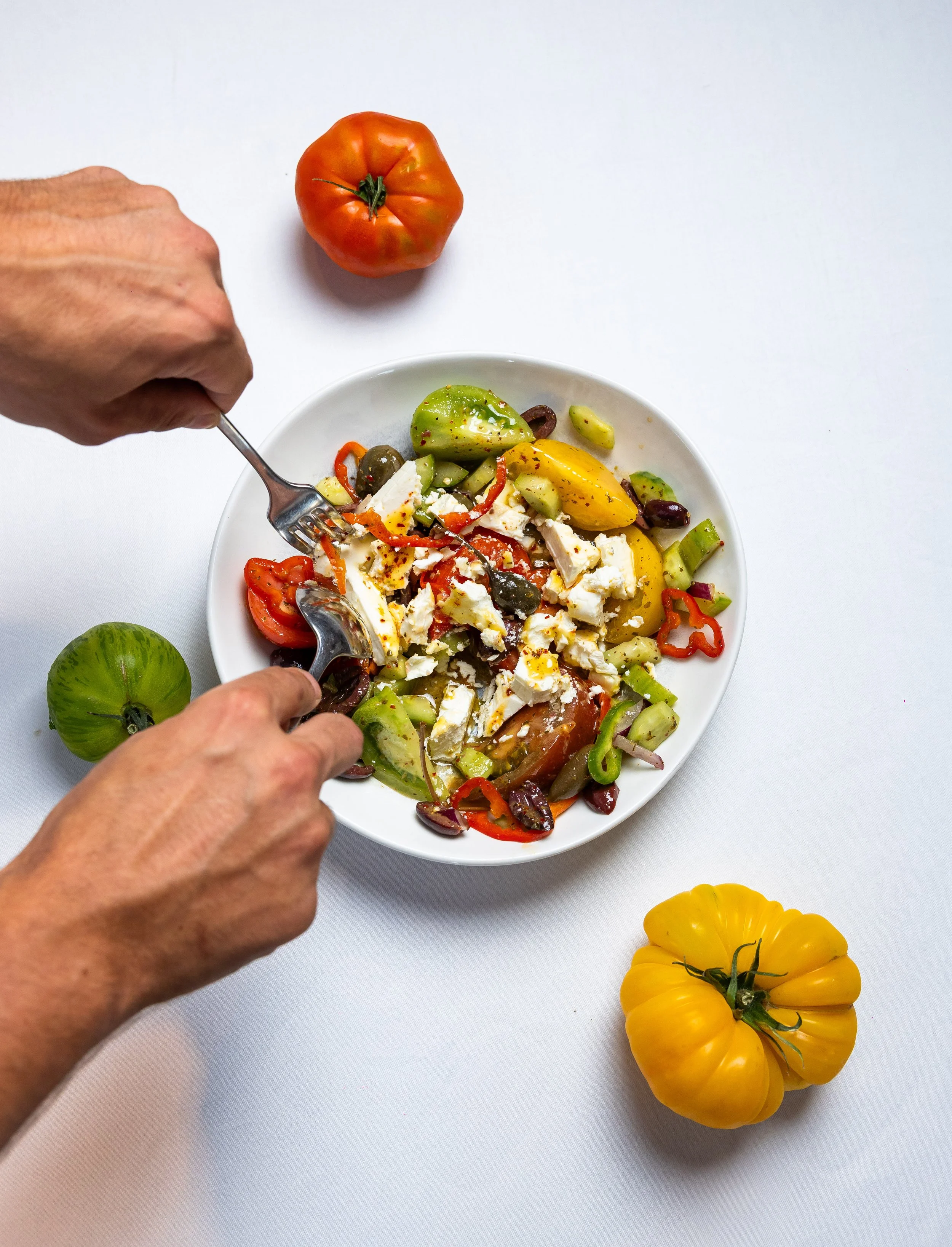 A white bowl of Greek salad with tomatoes, cucumbers, cheese, olives, and peppers on a white surface. A hand with a fork is mixing the salad. There are heirloom tomatoes on the surface, including a green tomato and a yellow tomato. A yellow heirloom 