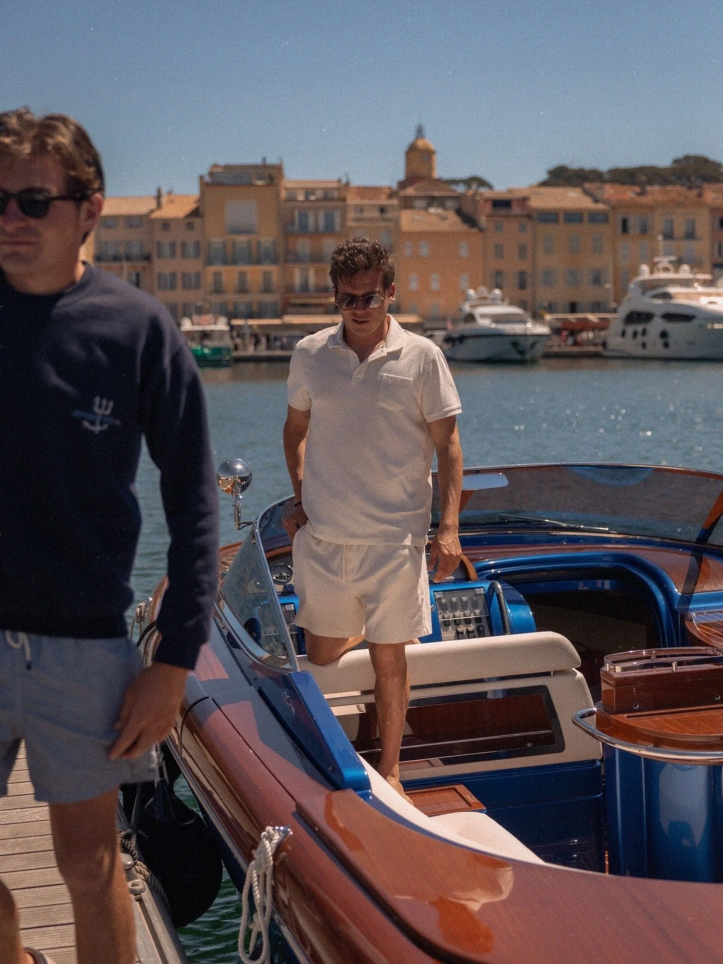 A man in white shorts and a white polo shirt standing on a boat docked by a marina with yachts and colorful mediterranean-style buildings in the background.