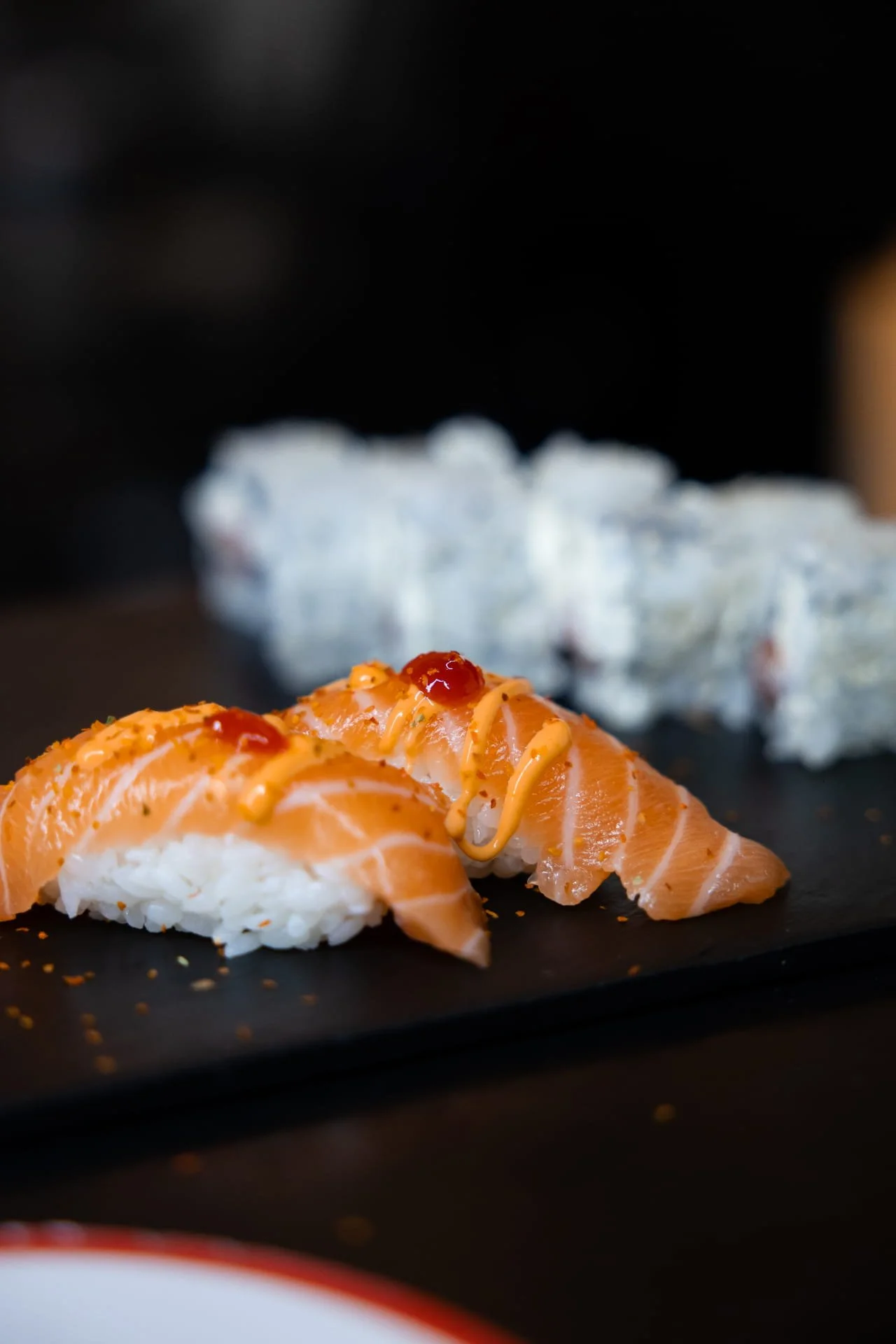Close-up of salmon sushi with sauce and garnishes on a black serving board.