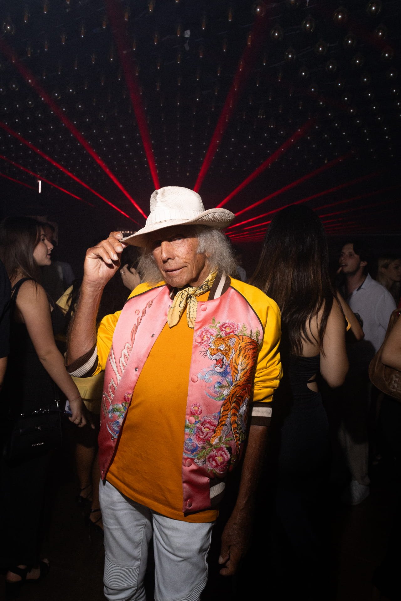 A man wearing a white cowboy hat, colorful satin jacket with embroidered tiger, orange shirt, and white pants, standing in a dark club with red laser lights and people in the background.