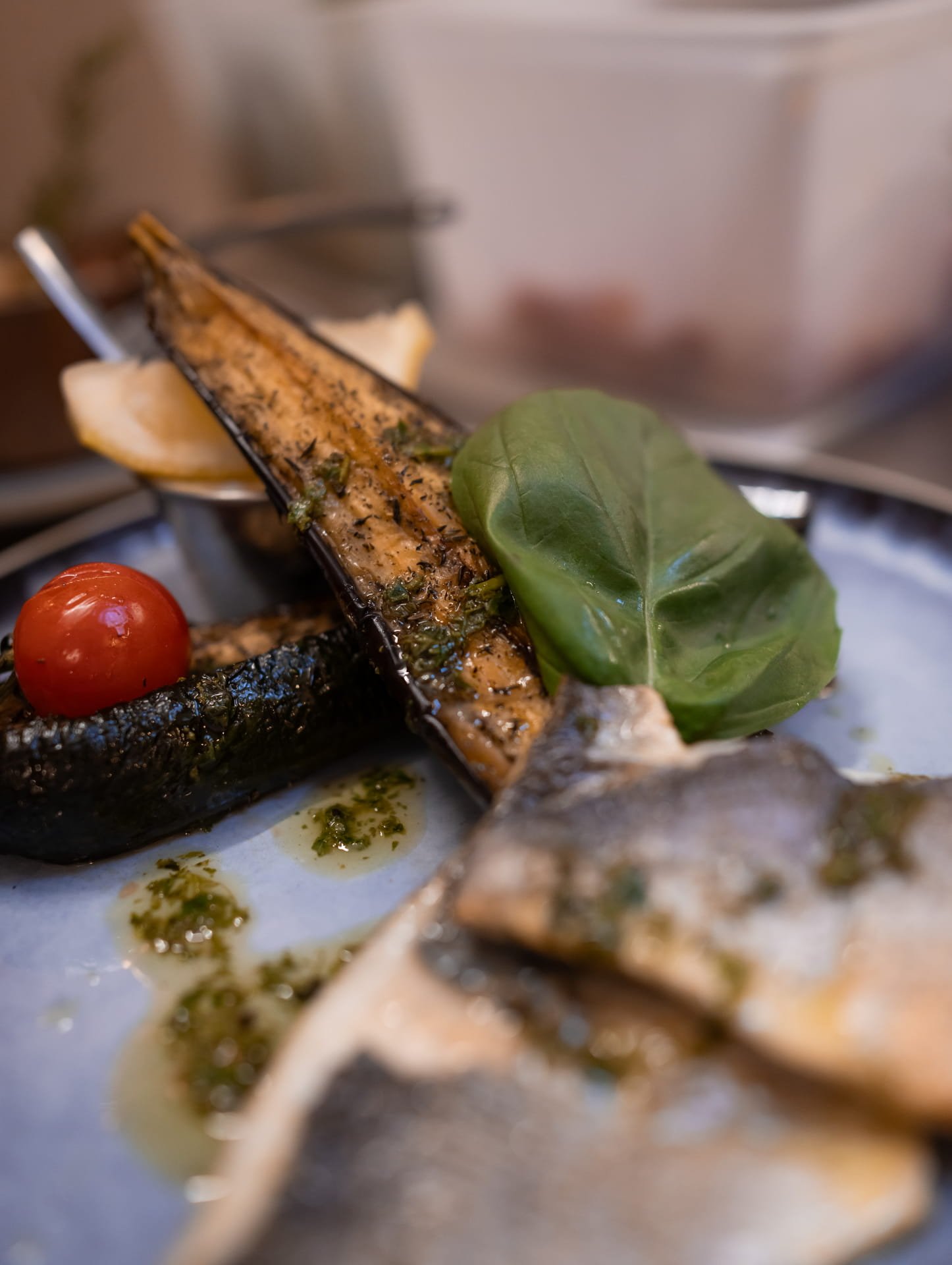 Close-up of grilled fish fillet garnished with a tomato and a basil leaf, with some herbs and sauce on the plate.