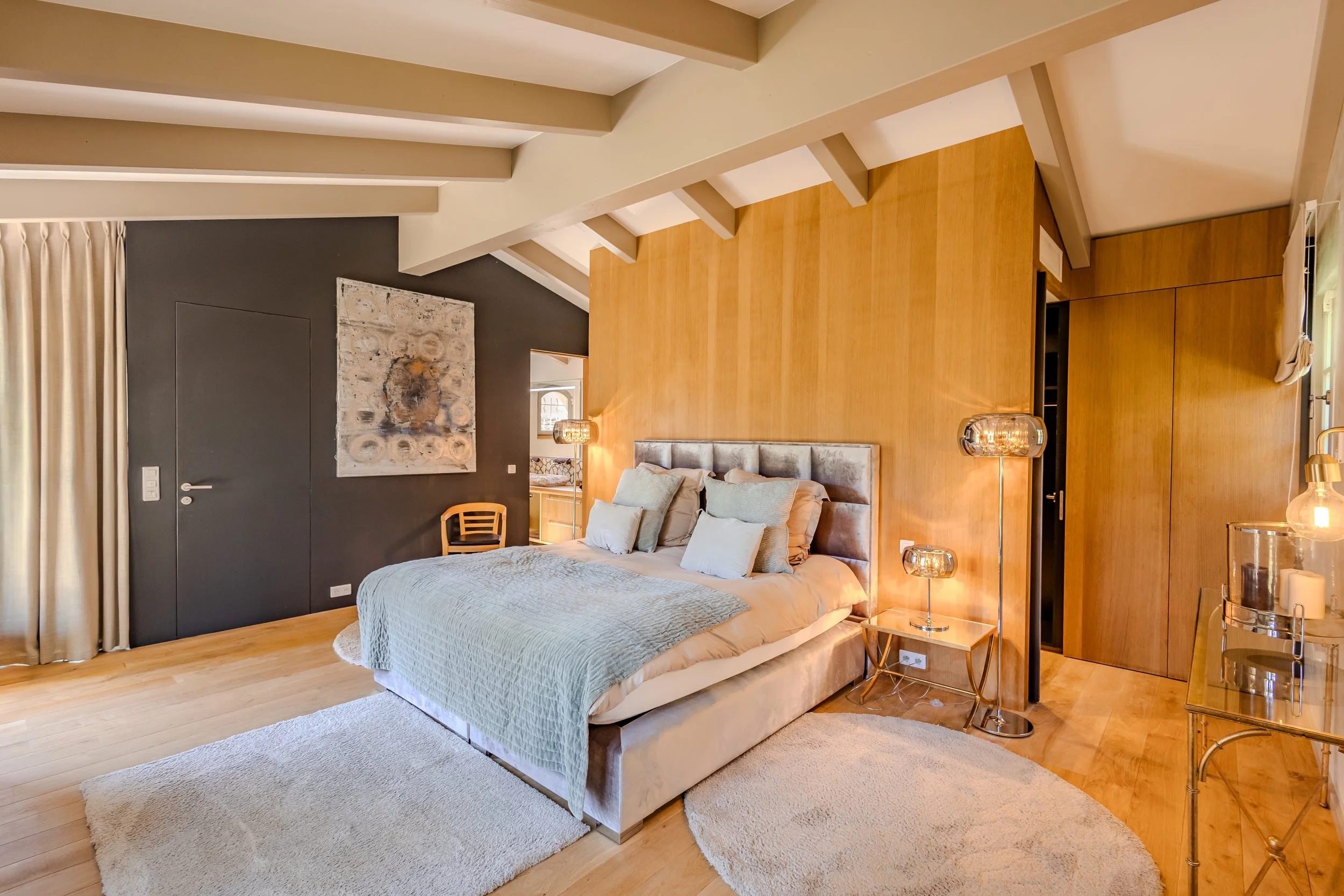 A cozy bedroom with a large bed, beige bedding, and several pillows. The room features wood-paneled and dark painted walls, a soft area rug, and stylish lighting fixtures. There's a small wooden chair and modern decor, including a painting and a mirr