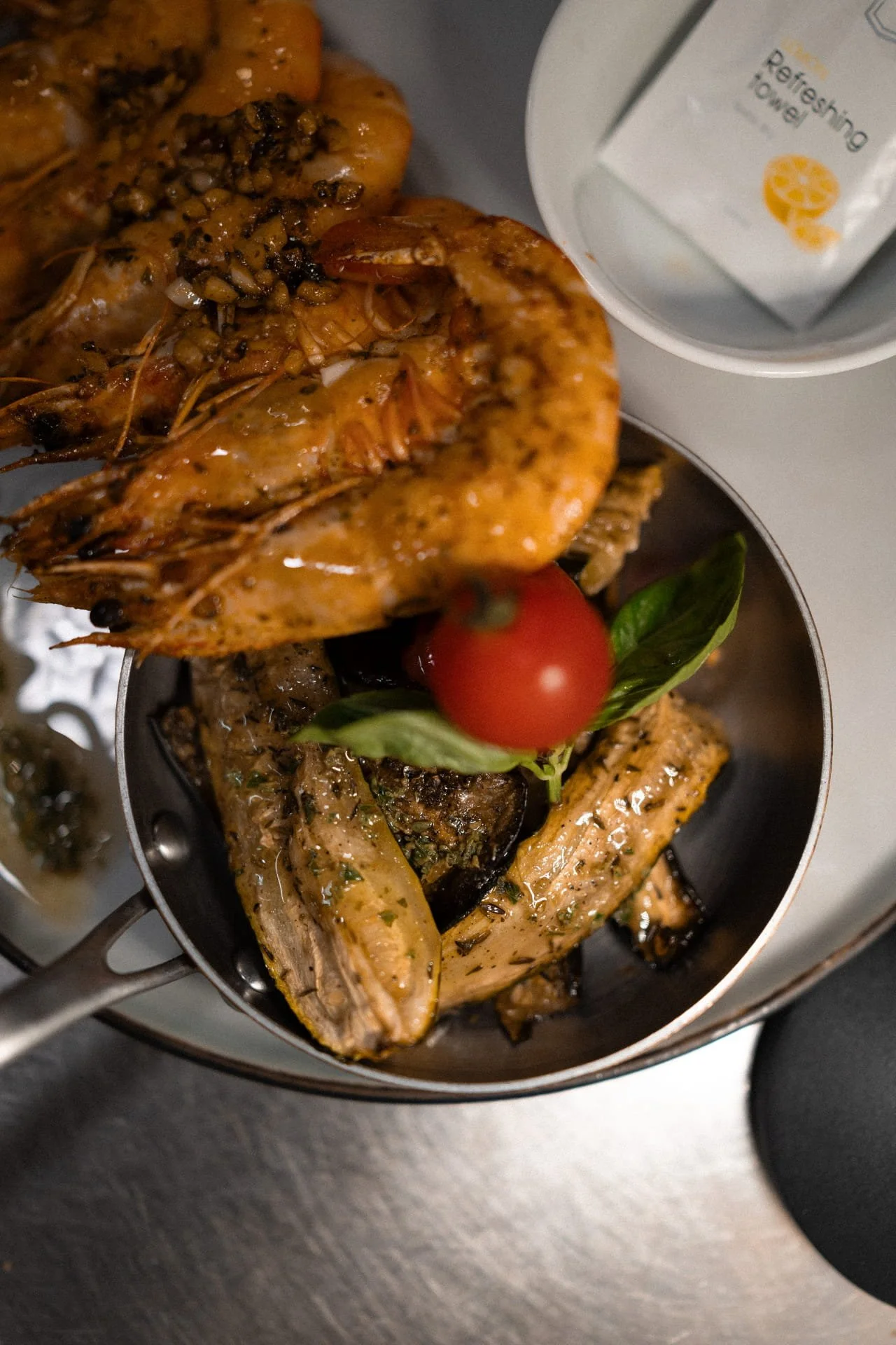 Close-up of cooked shrimp topped with garlic and herbs, garnished with a cherry tomato and basil leaf, in a small skillet. Part of a white bowl with paper inside is visible in the background.