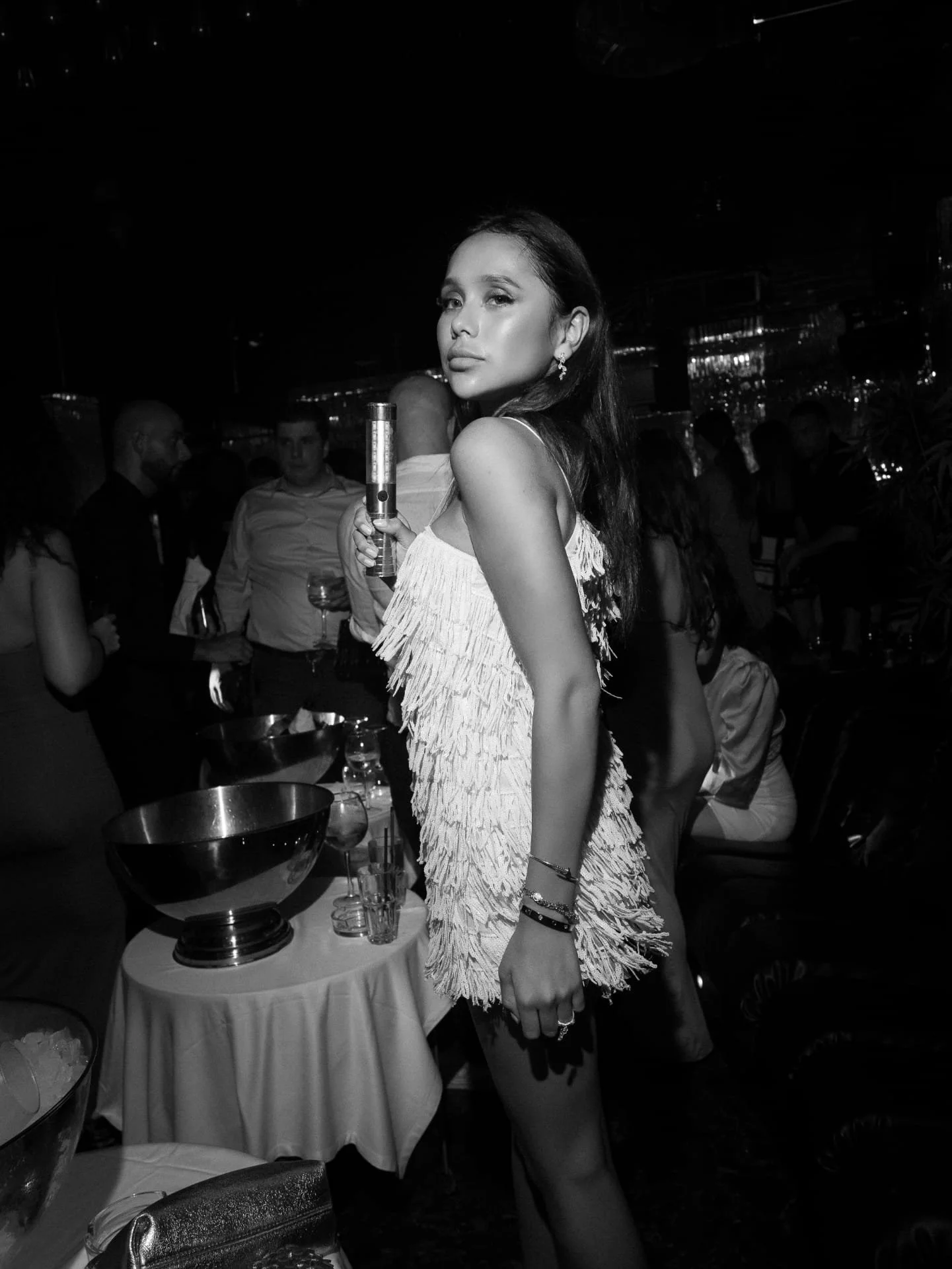 Black and white photo of a young woman with dark hair posing at a social event. She is holding a small flashlight and wearing a fringed dress, with a crowd of people in the background.