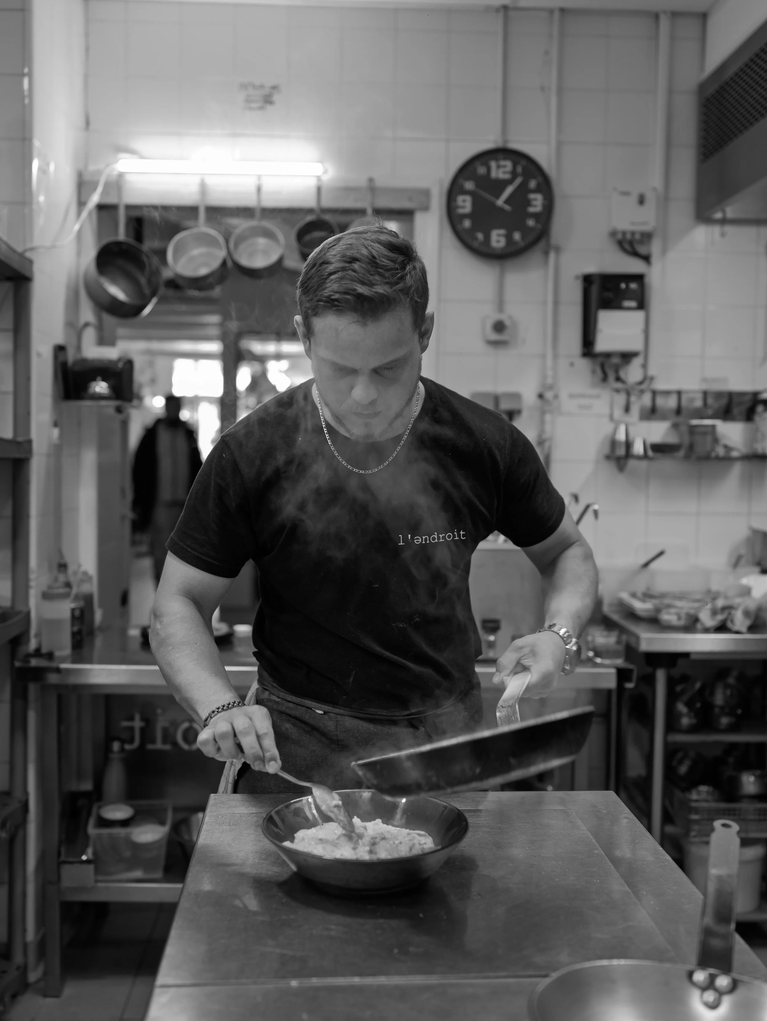 A man with short hair wearing a black t-shirt and jewelry is standing in a kitchen, pouring food from a pan into a bowl on the table.