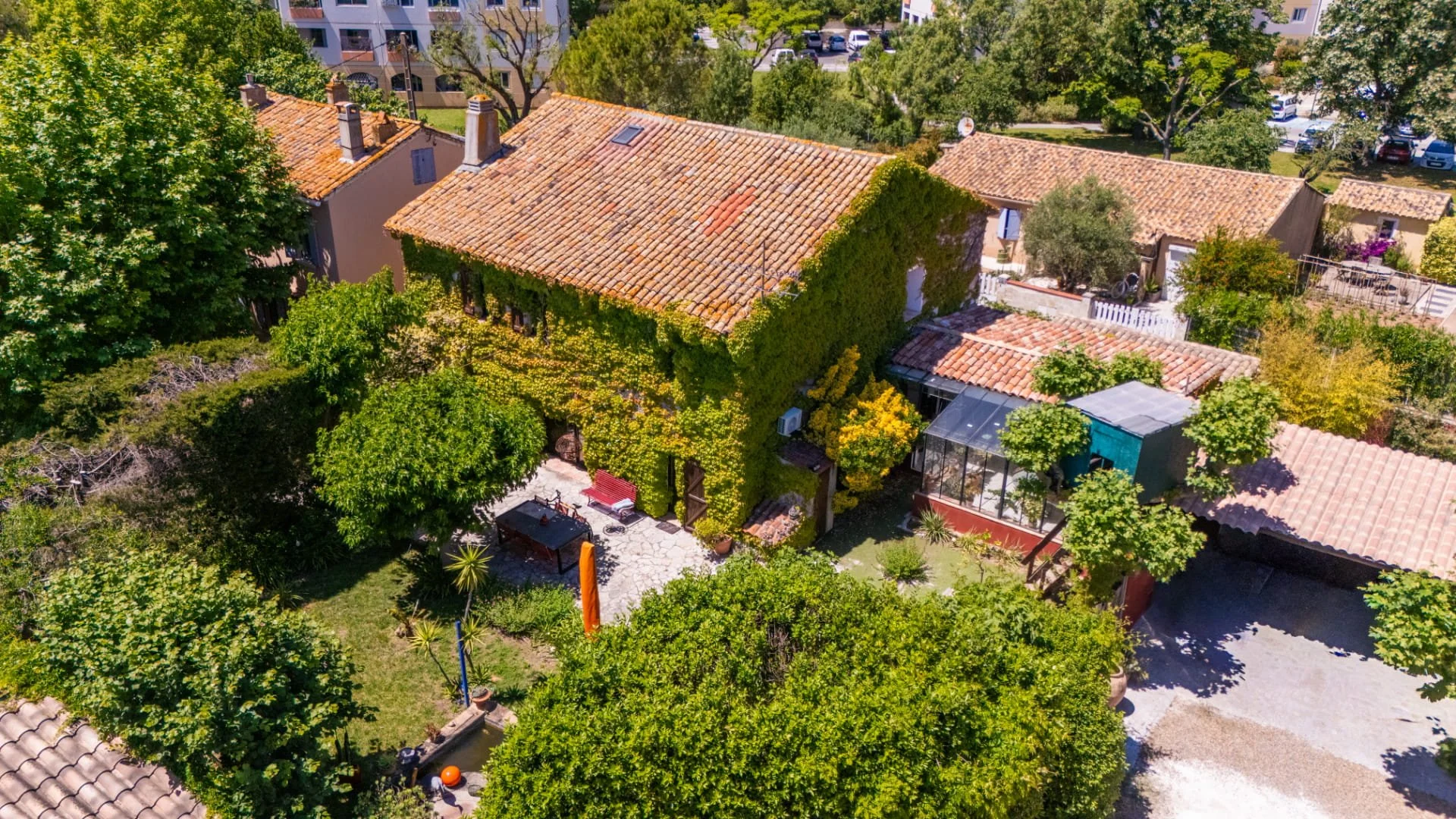 Aerial view of a residential property with tiled roofs, lush green trees, a garden area, and a small patio with outdoor furniture.