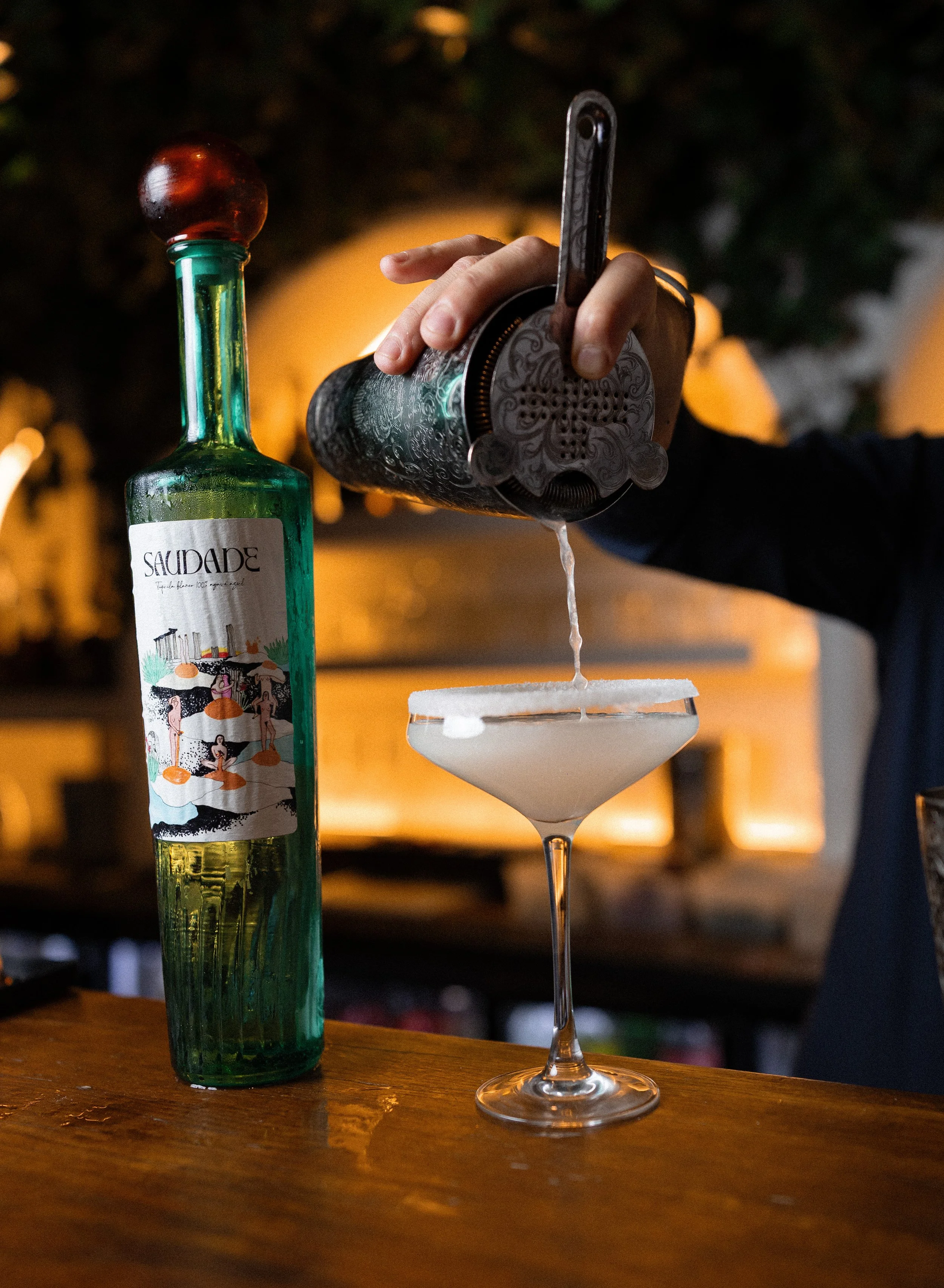 A hand pours liquid from a vintage metal cocktail shaker into a for a cocktail glass on a wooden table with a bottle of Sauza tequila nearby.