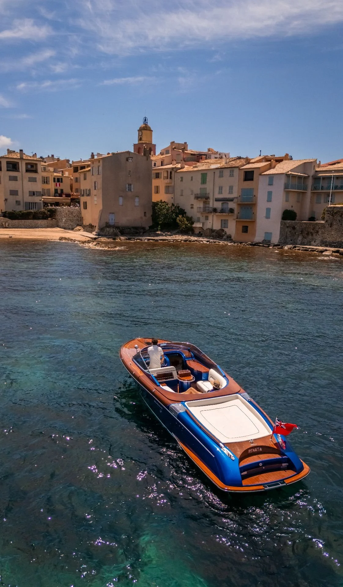 A boat on calm blue water near a shore with a row of multi-story buildings and a tower in the background under a partly cloudy sky.
