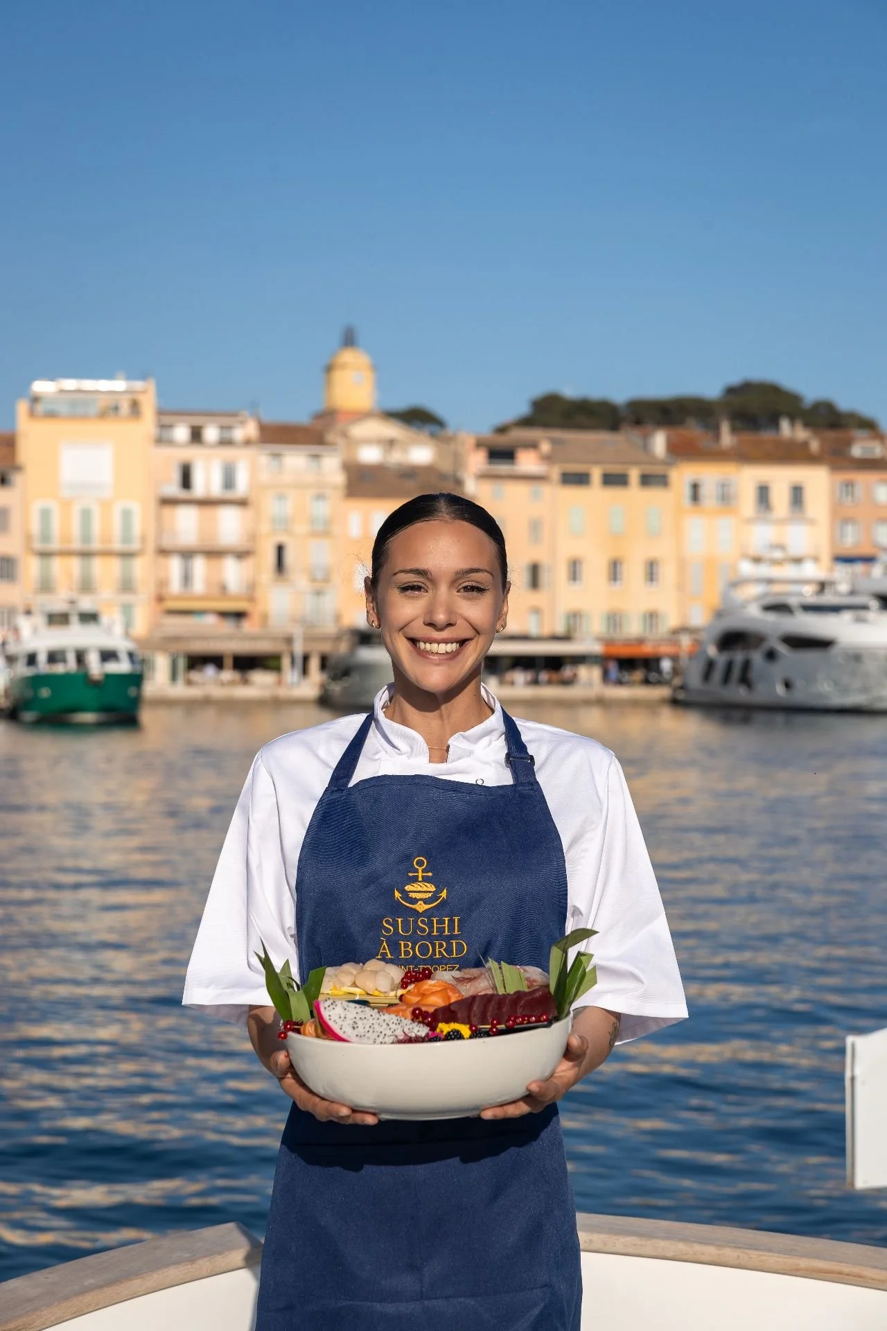 A woman in a white chef's jacket and navy apron holding a large bowl of sushi, standing outdoors near water with buildings and boats in the background.
